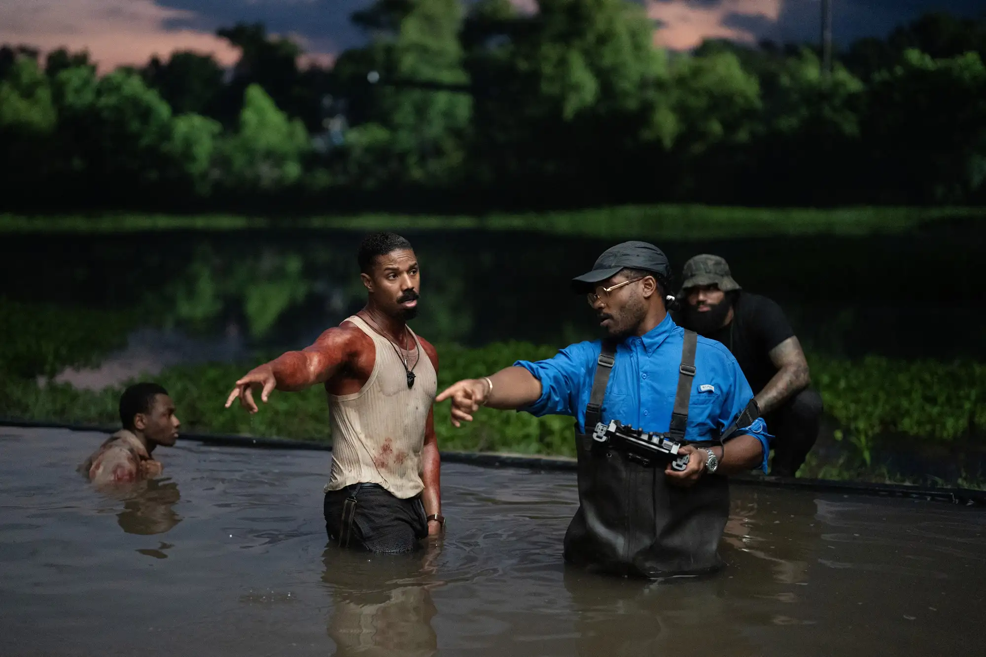 Four men standing and wading in a body of water near lush vegetation at dusk, with two of them pointing in opposite directions.