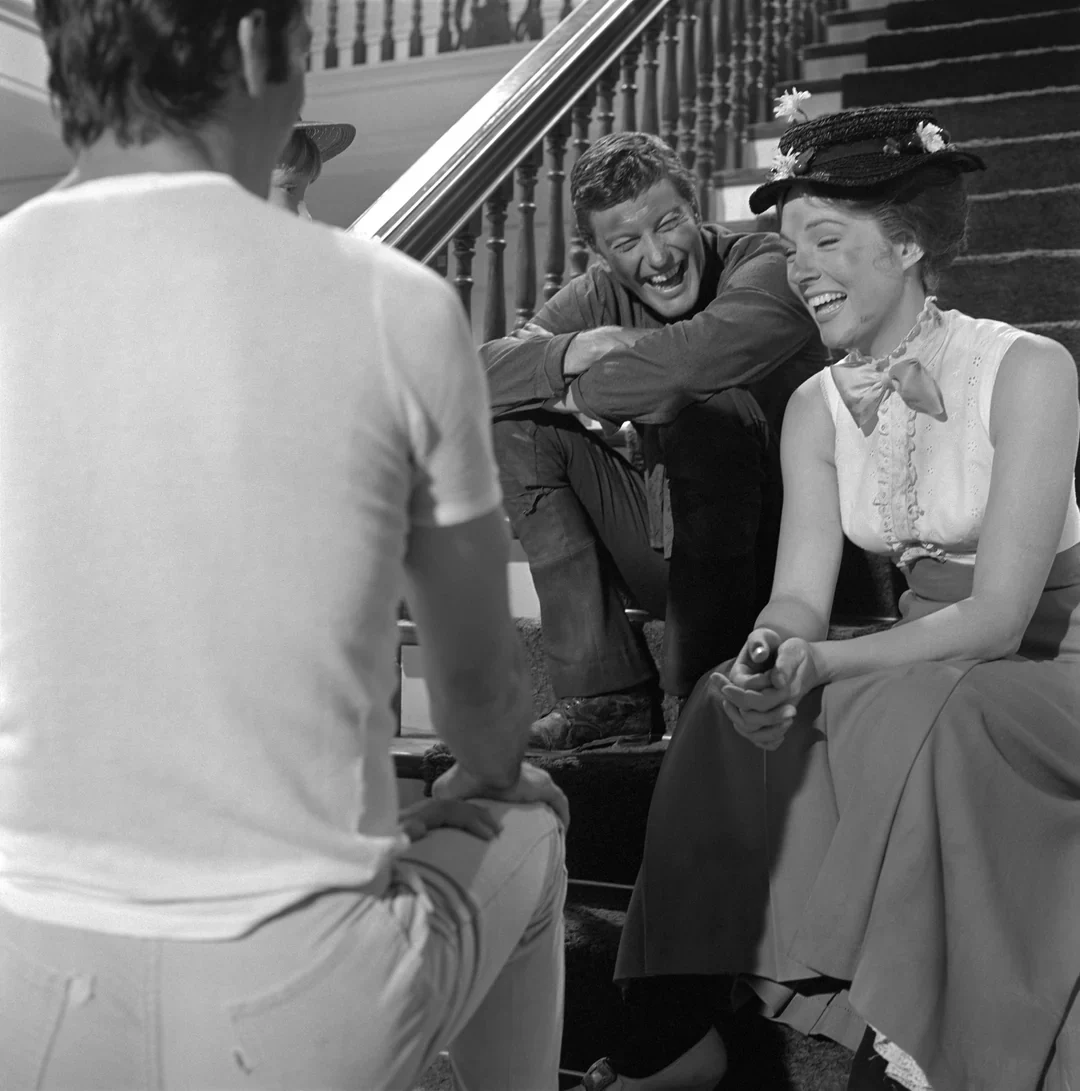 A woman with a hat and bow tie and a man sitting on stairs laughing, while a man with his back to the camera sits in front of them.