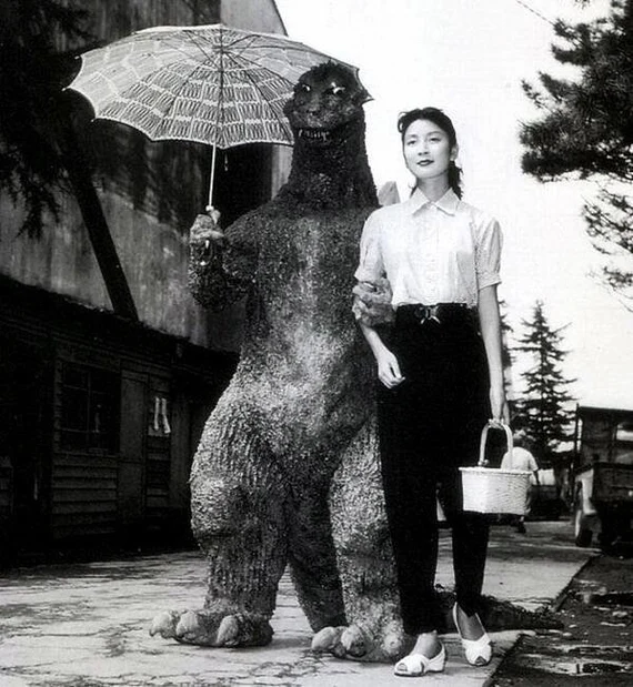 Black and white photo of a woman walking arm-in-arm with a person in a Godzilla costume holding an umbrella.