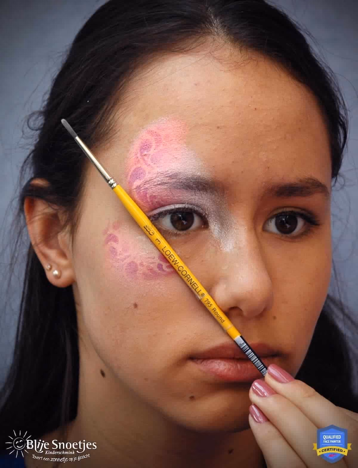 a woman with a fancy eye face paint design being made holding a brush demonstrating to the focal point