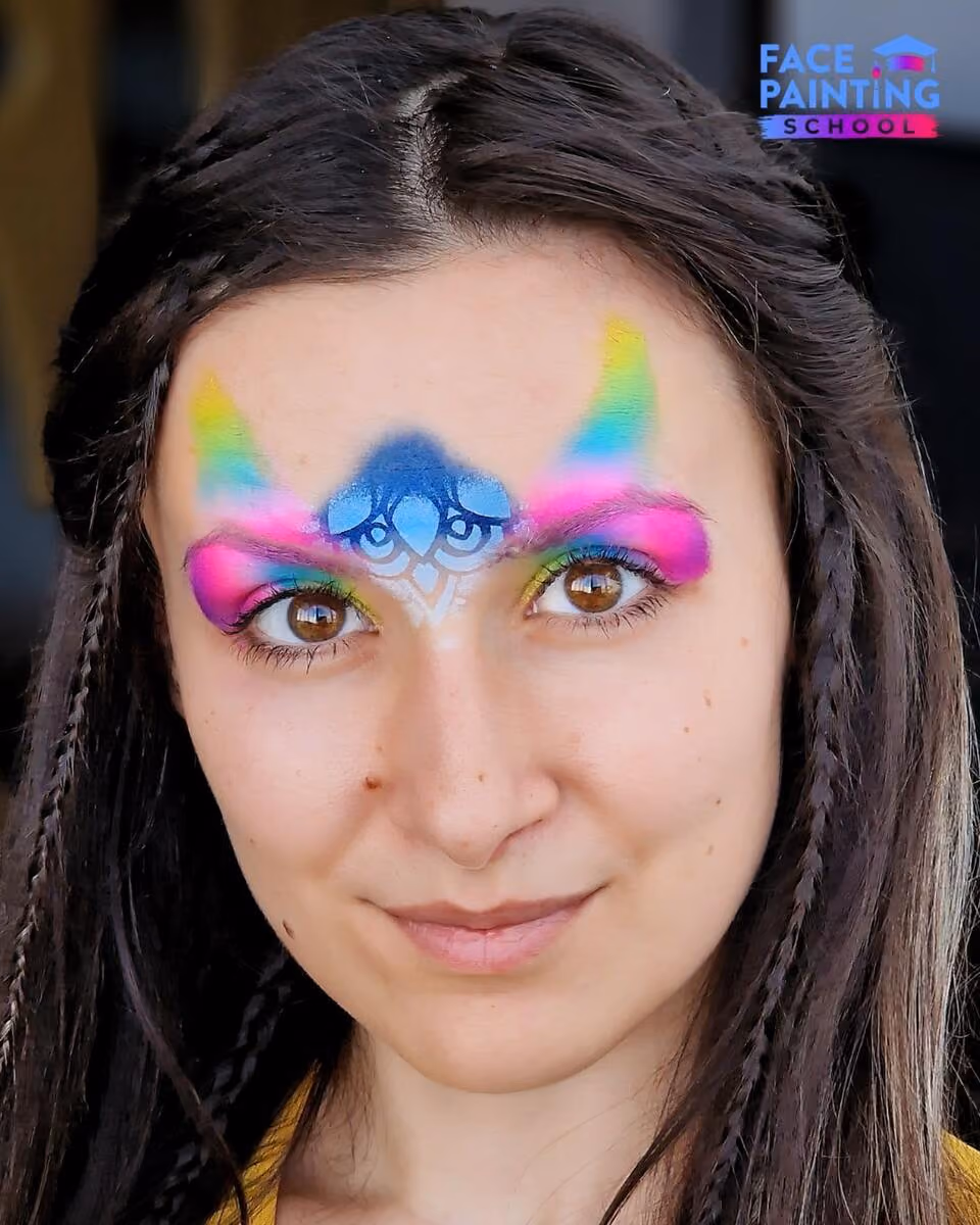 a woman with a shiny rainbow colored paint over her eyes with a blue beginning of a flower on her forehead