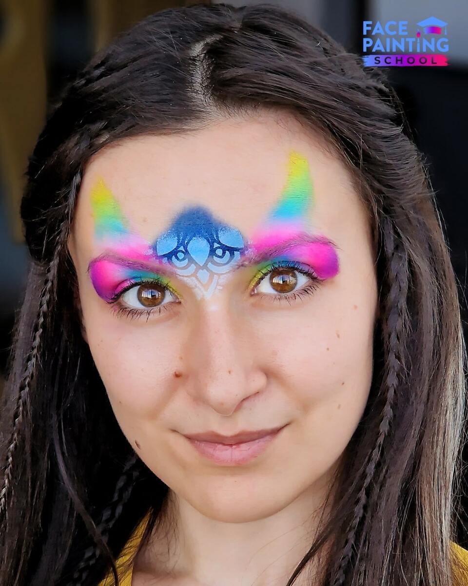 a woman with a shiny rainbow colored paint over her eyes with a blue beginning of a flower on her forehead
