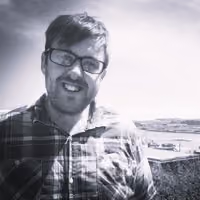 Man wearing glasses and a plaid shirt smiling outdoors with a landscape and partly cloudy sky in the background.