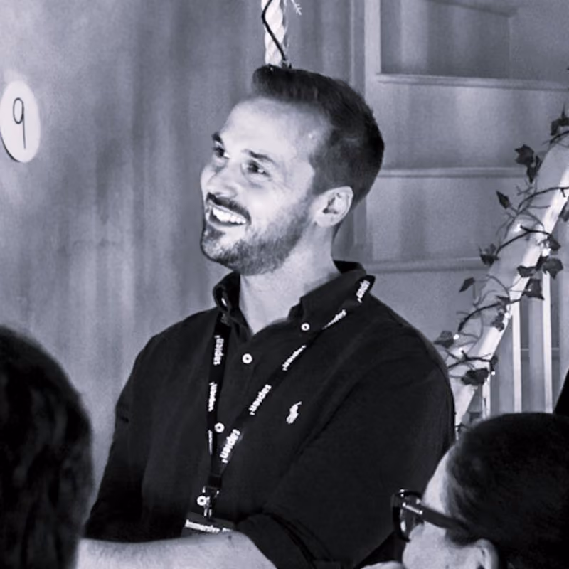 Smiling man with a beard and short hair wearing a black polo shirt and lanyard, engaged in conversation indoors.