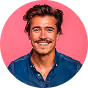 Smiling young man with brown hair and mustache wearing a blue shirt against a pink circular background.