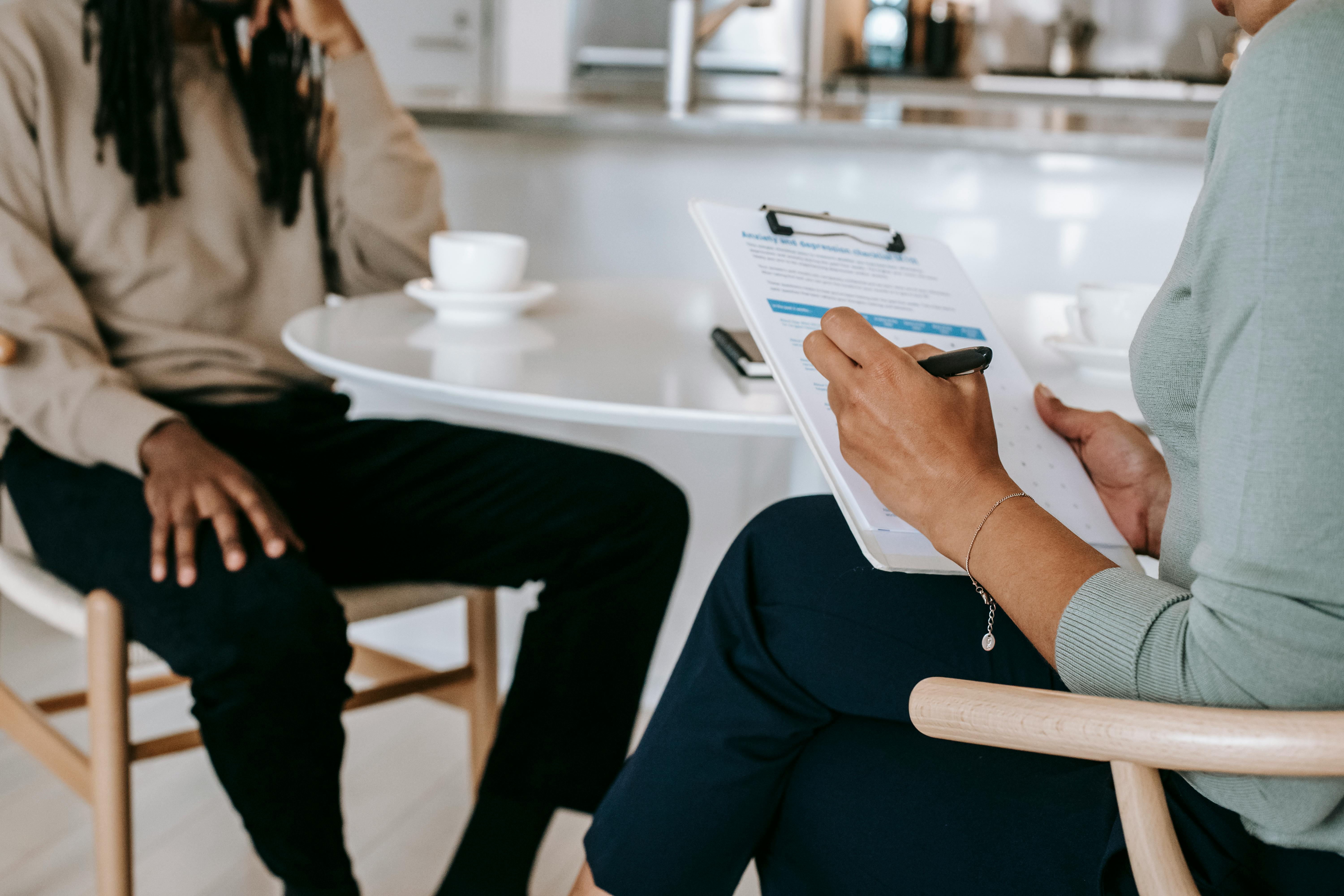 A person holding a clipboard and pen interviewing another person seated at a round table with coffee cups.