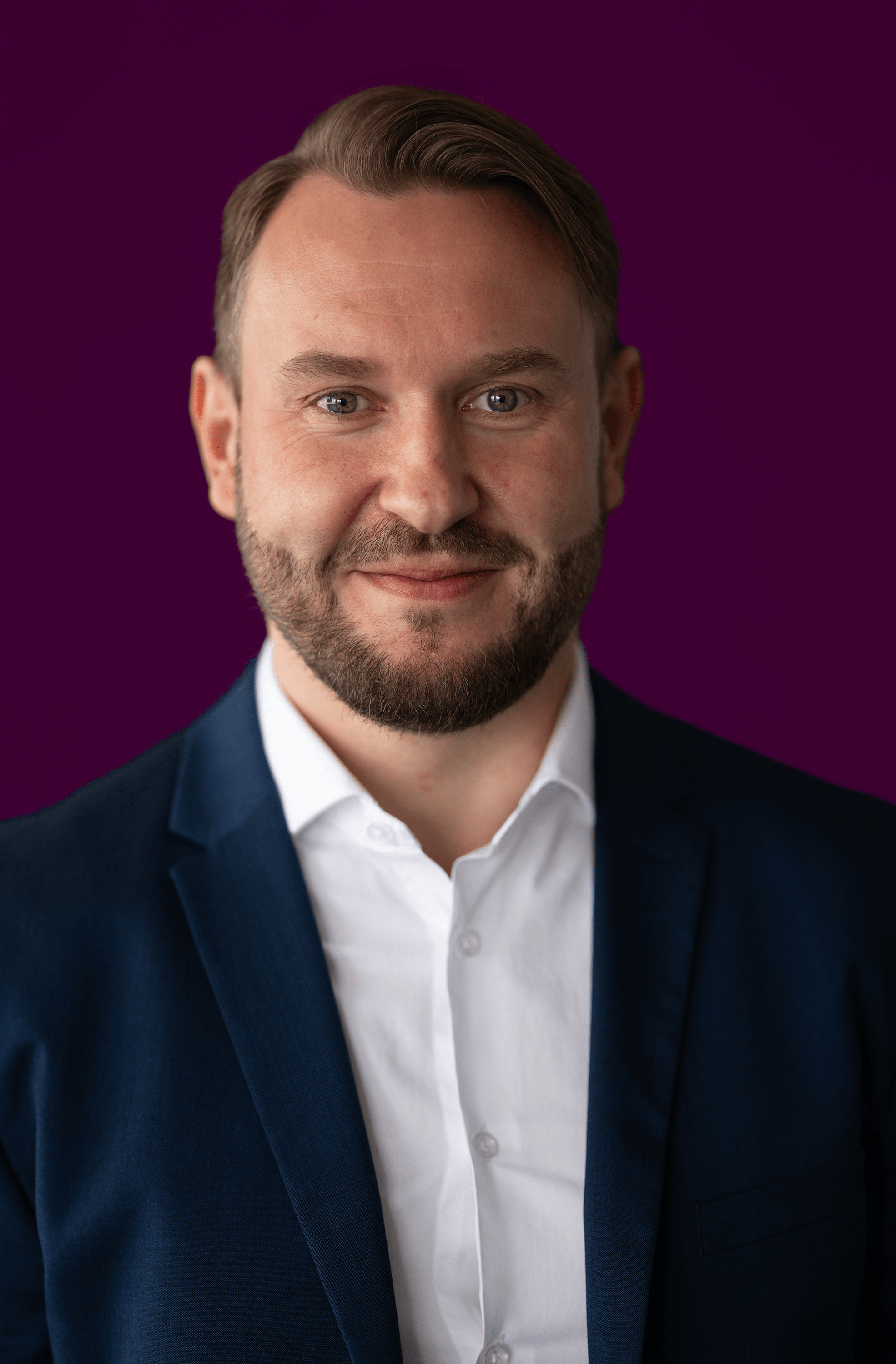 Man with short brown hair and beard wearing a white shirt and navy blazer against a plum purple background.