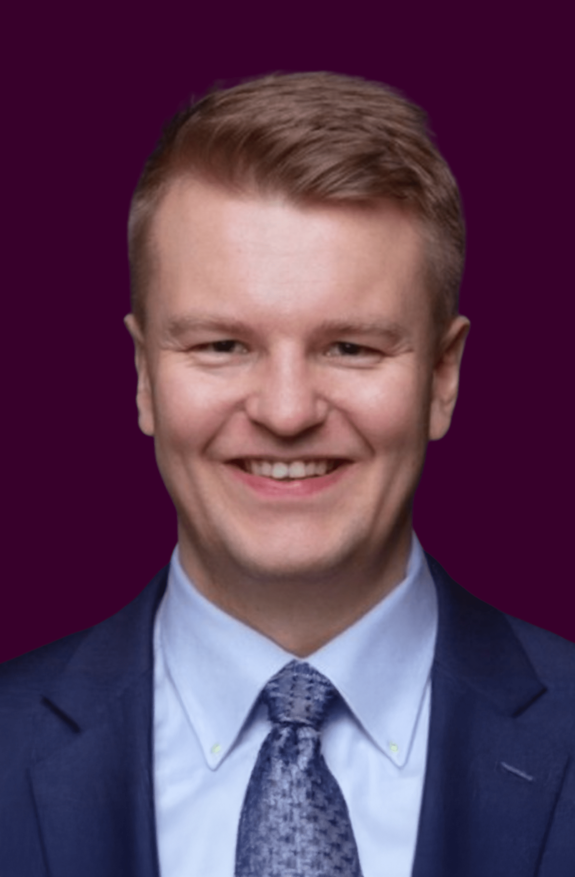 Smiling man with short light brown hair wearing a blue suit, light blue shirt, and patterned tie against a dark purple background.