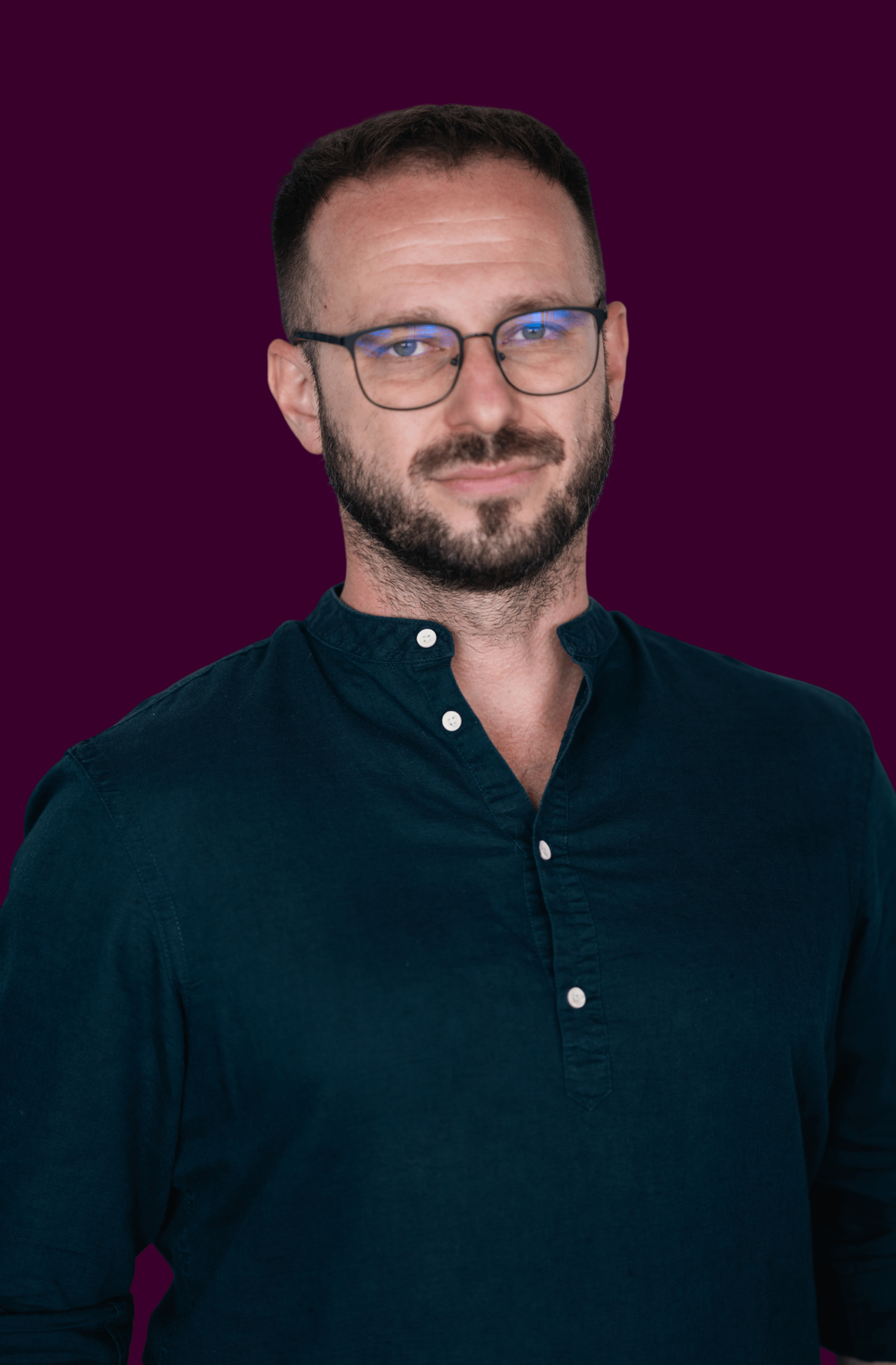 Man with short hair, beard, and glasses wearing a dark blue buttoned shirt against a purple background.