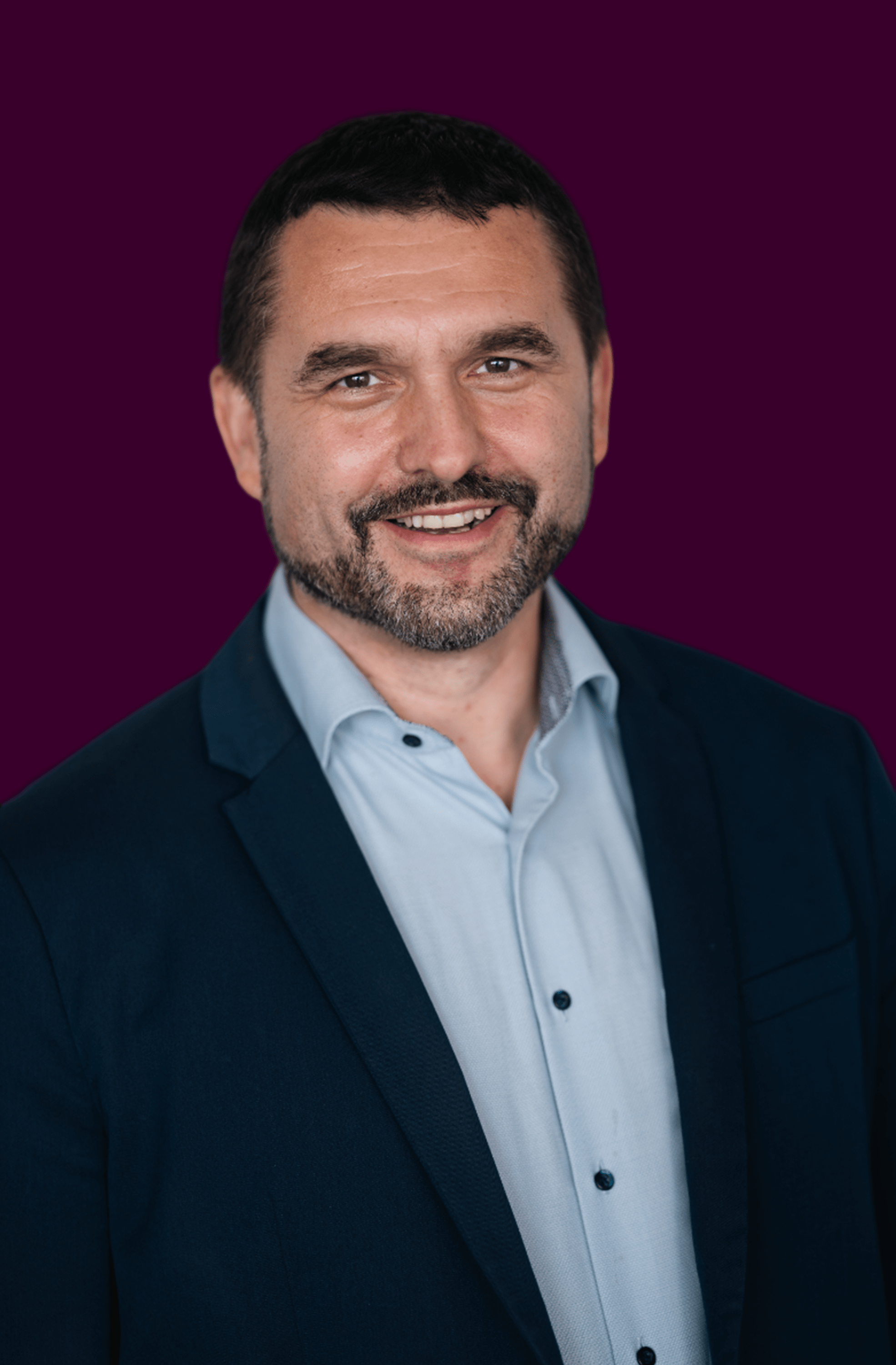 Smiling man with short dark hair and beard wearing a light blue shirt and dark blazer against a dark purple background.