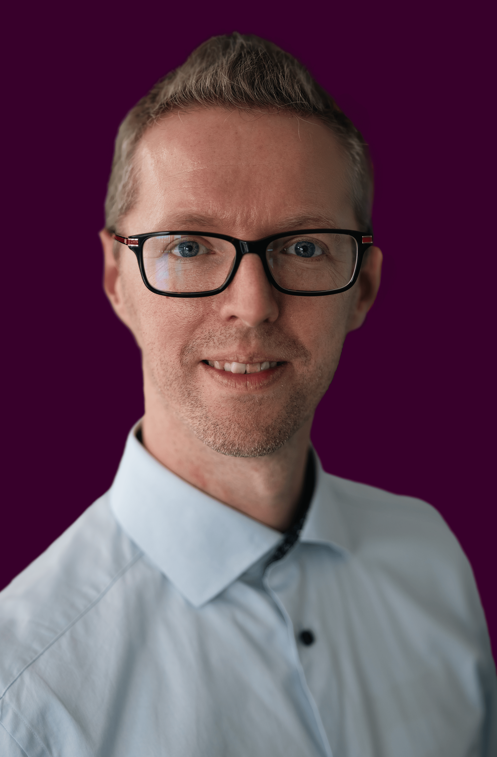 Smiling man with short light brown hair, glasses, and a light blue shirt against a dark purple background.