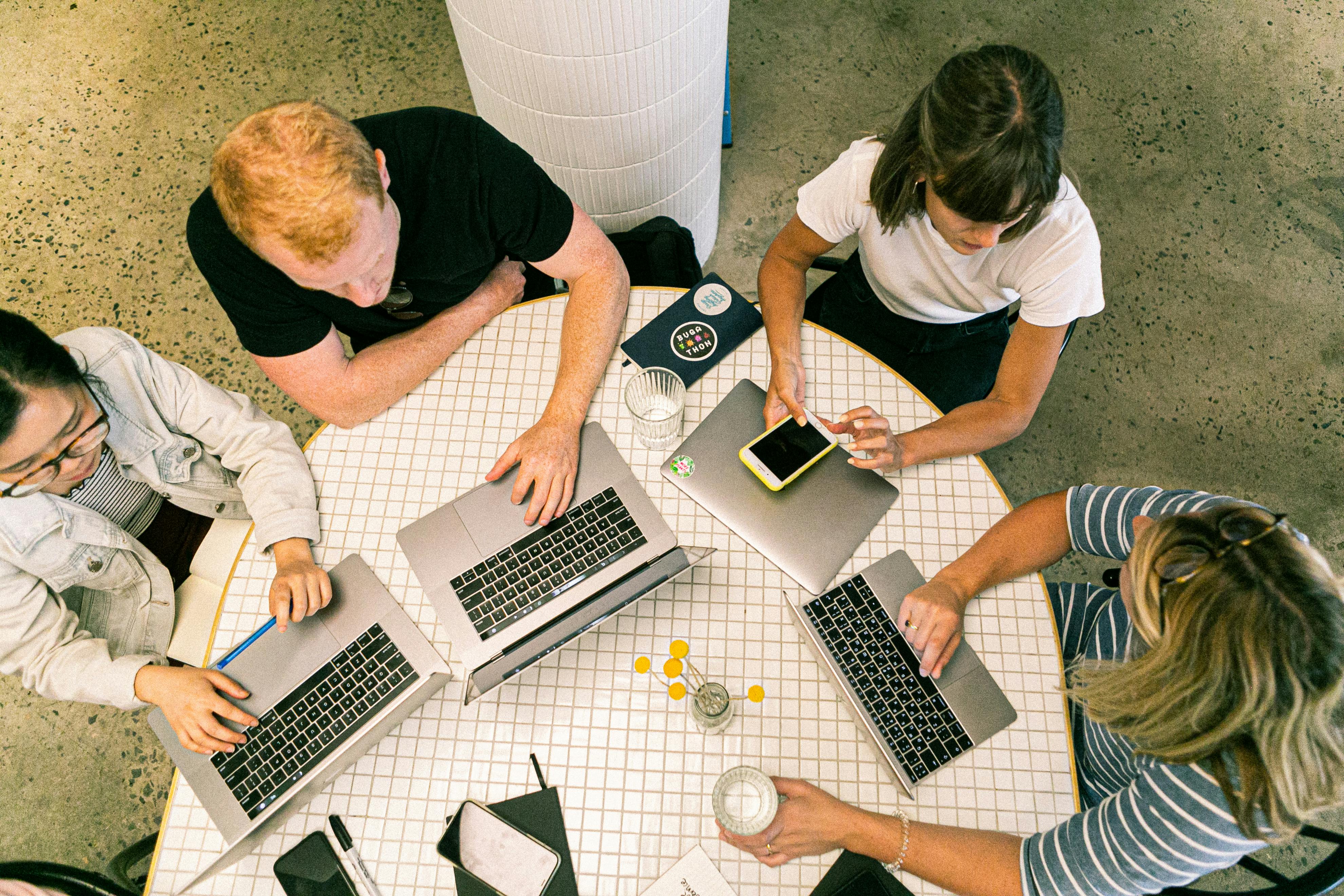Four people sitting around a round table working on laptops and a smartphone in a collaborative setting.