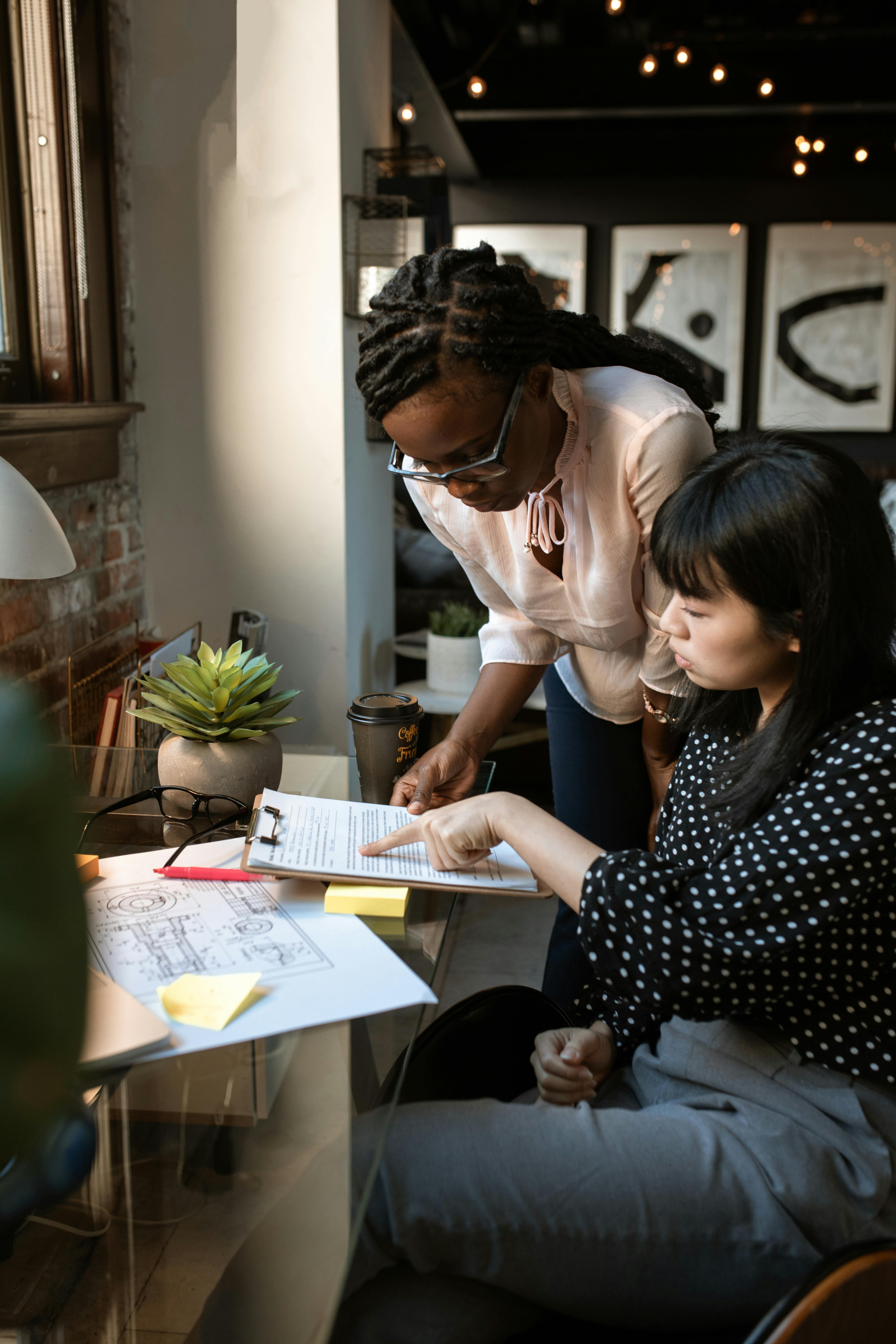 Two women working together at a desk reviewing a document on a clipboard in a modern office setting.