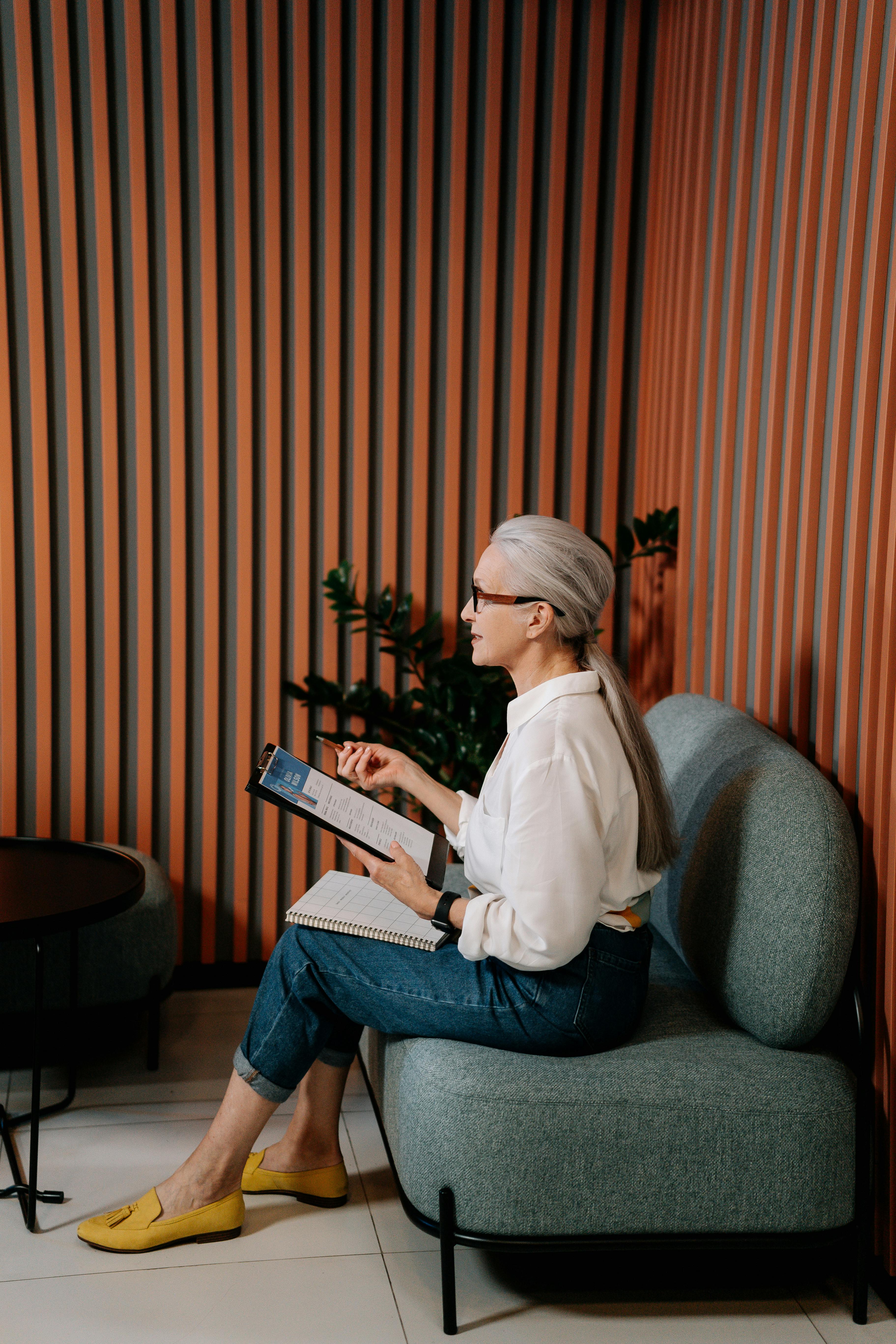 Elderly woman with gray hair tied back sitting on a gray cushioned sofa, holding a clipboard and pen, wearing glasses, white shirt, blue jeans, and yellow shoes.