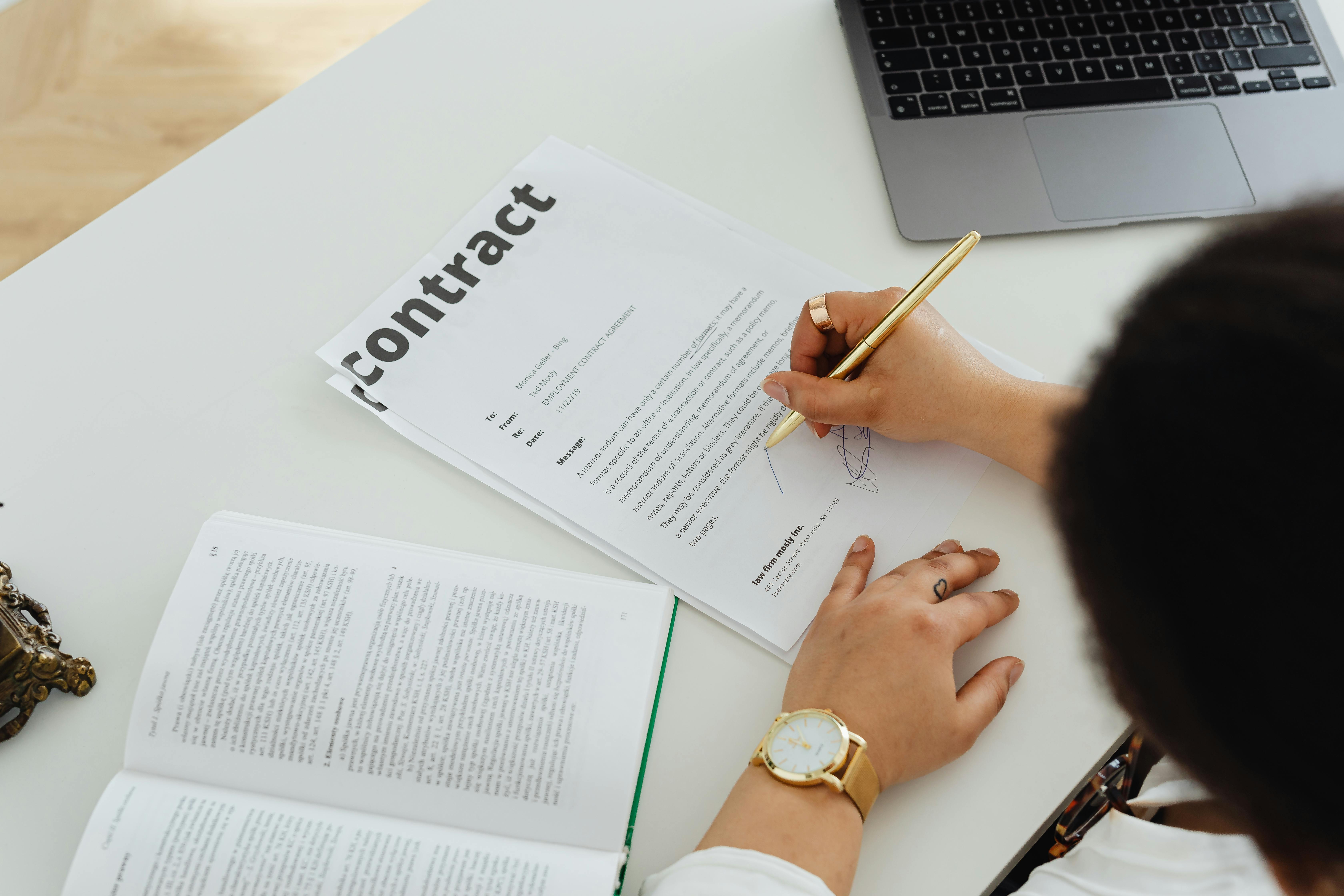 Person signing a contract on a white desk with an open book and a laptop nearby.