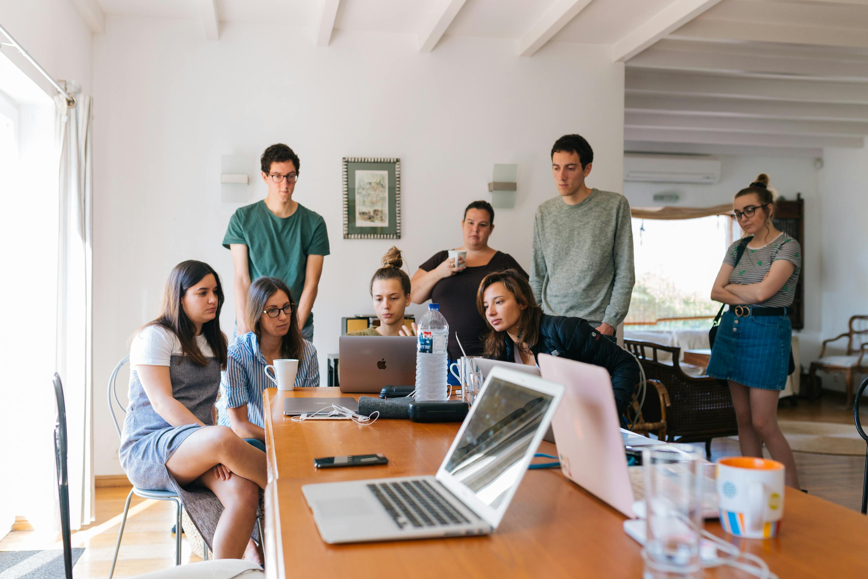 Group of eight young adults gathered around a wooden table watching laptop screens in a bright room.