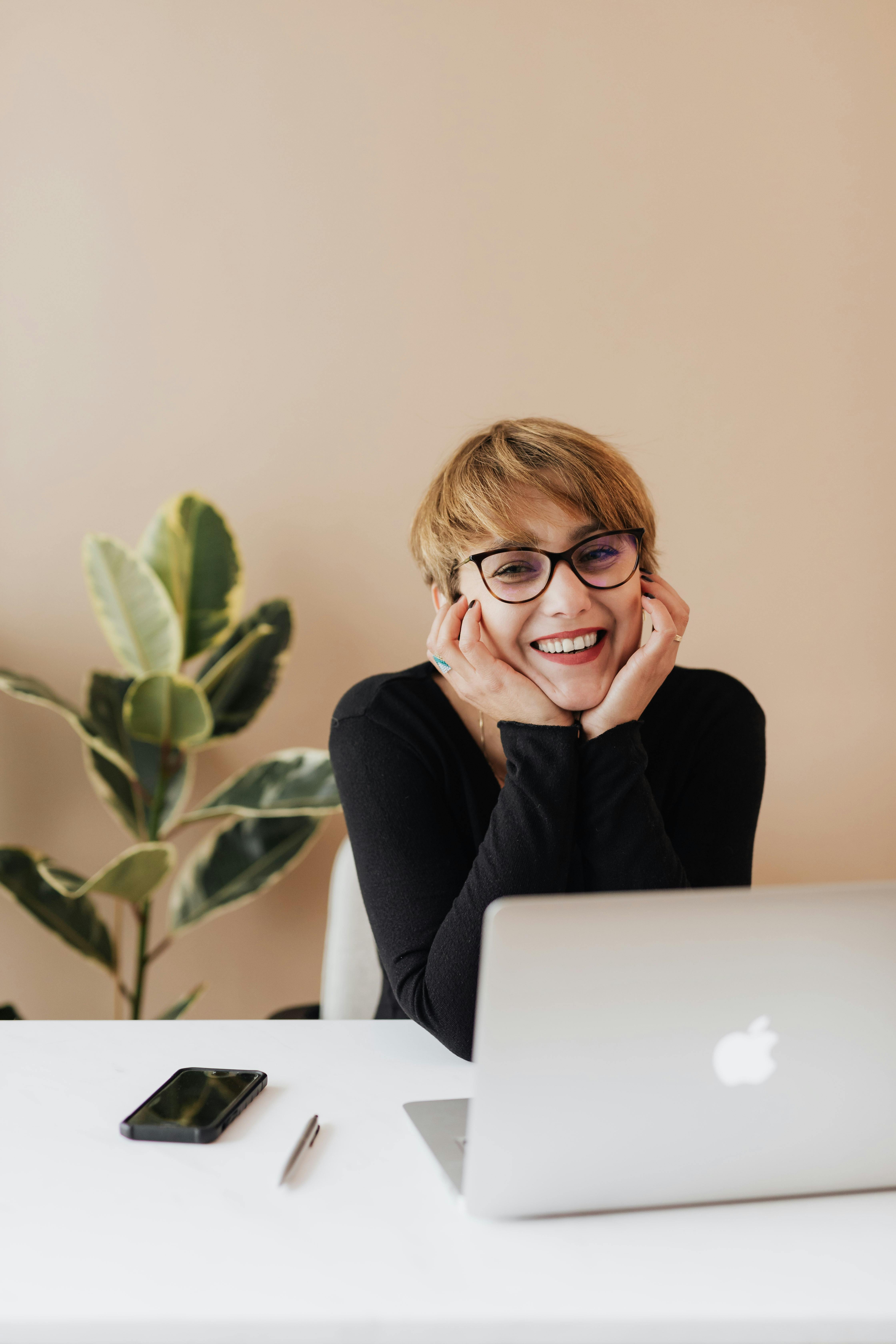 Smiling woman with glasses sitting at a desk with a laptop, smartphone, and pen, with a potted plant in the background.