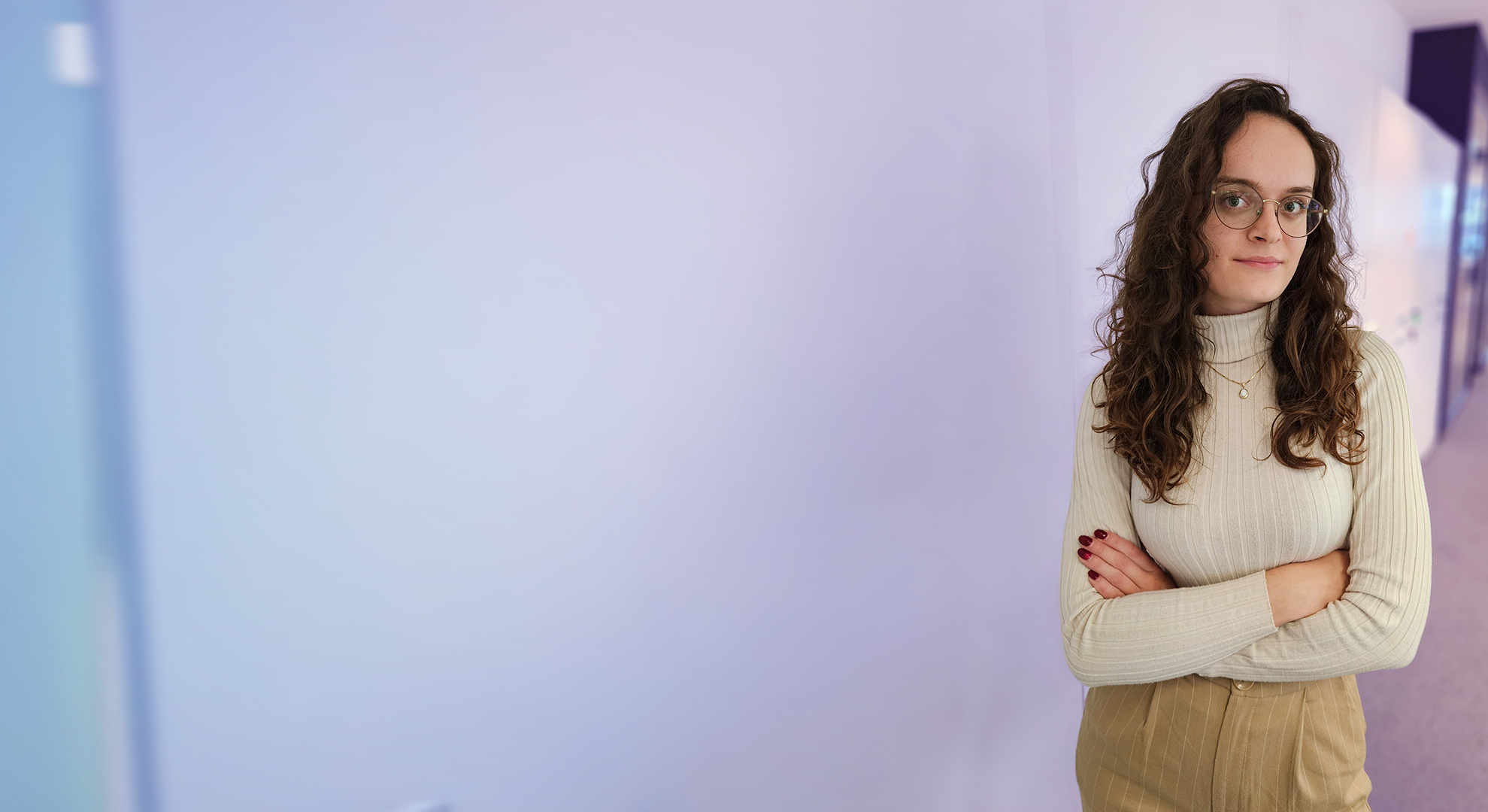 Young woman with long curly hair and glasses, wearing a beige turtleneck sweater and tan pants, standing with arms crossed in a softly lit hallway.