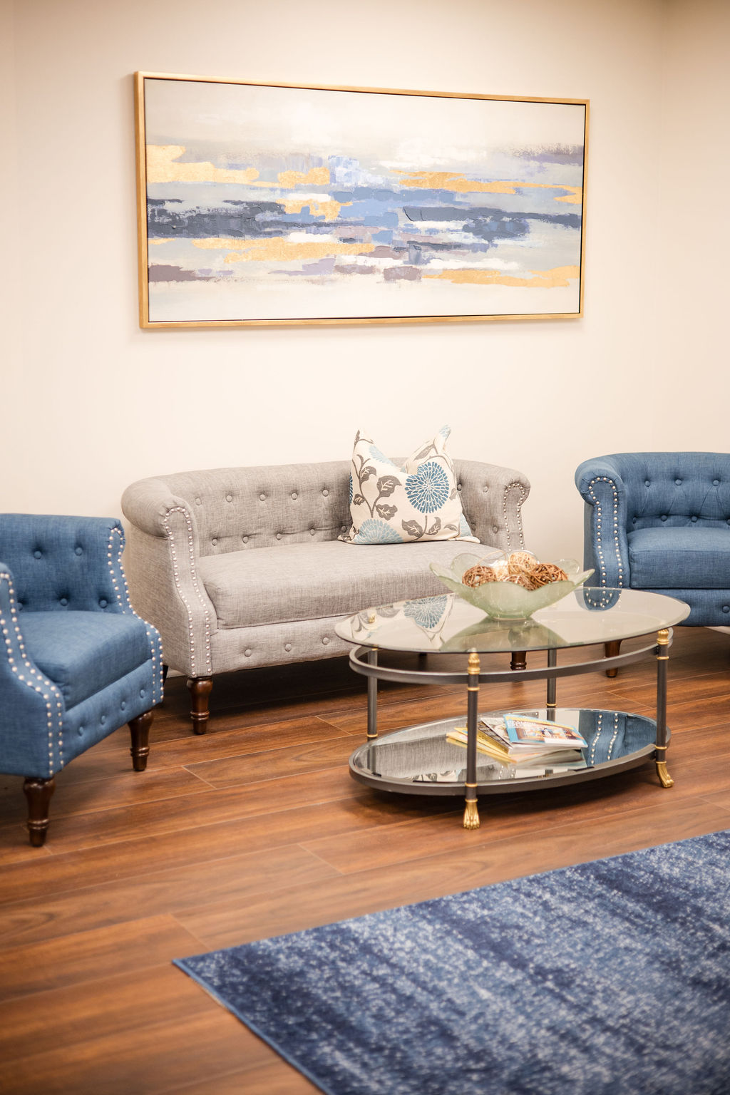Interior waiting room of the practice with a gray tufted loveseat, two blue armchairs with nailhead trim, glass coffee table with decorative bowl, and abstract painting on wall.