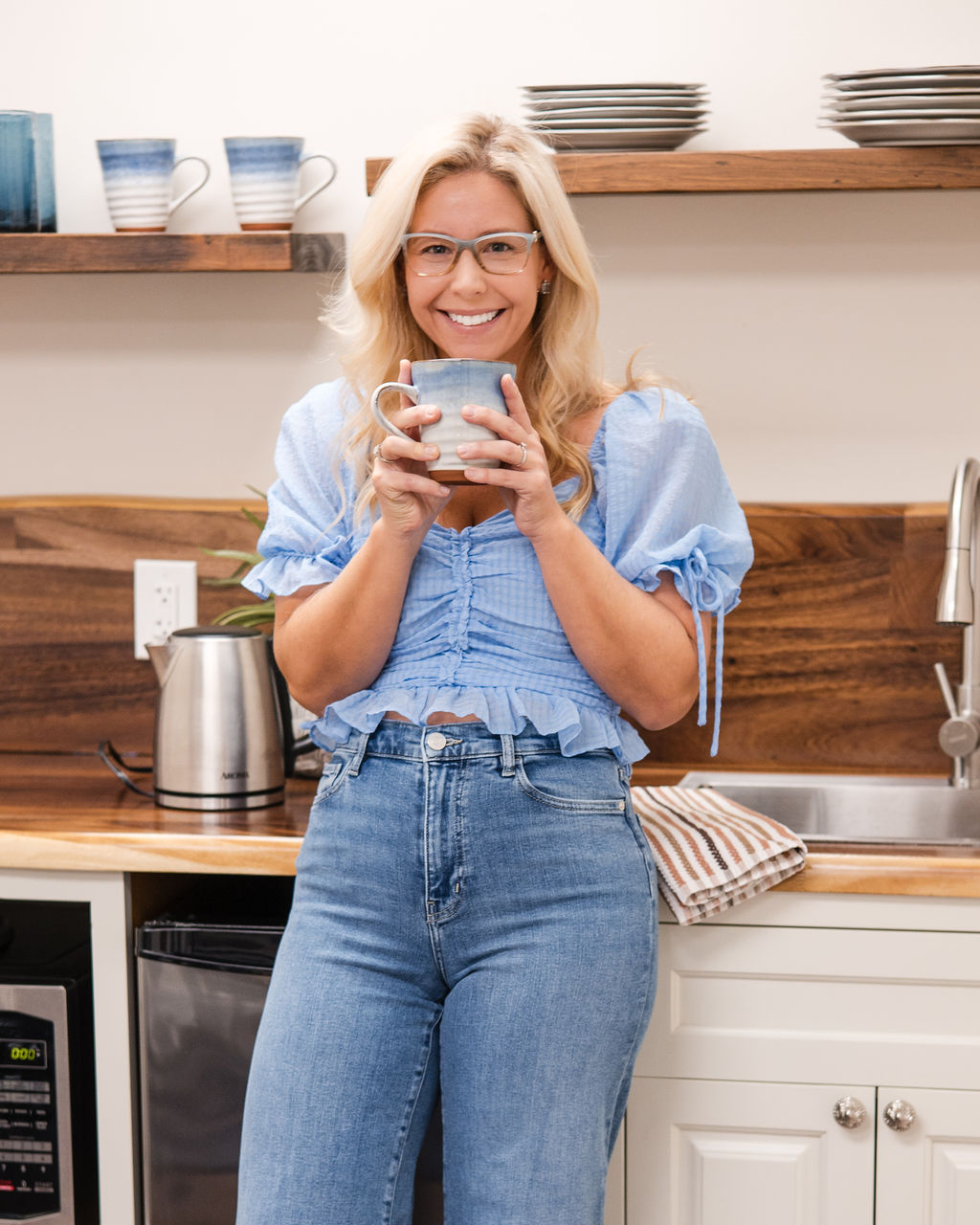 Lindsey Capelli, therapist and owner of, Living Well Counseling Center, in glasses and a light blue top holding a blue and white ceramic mug in a kitchen.