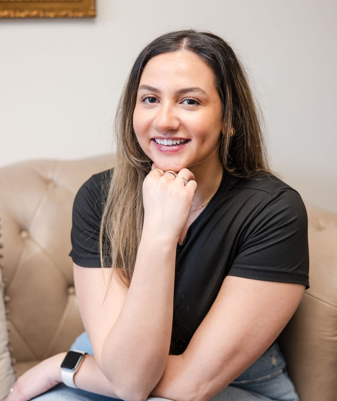 Carly Frascino, counselor provider, with long hair sitting on a beige tufted sofa, resting her chin on her hand.