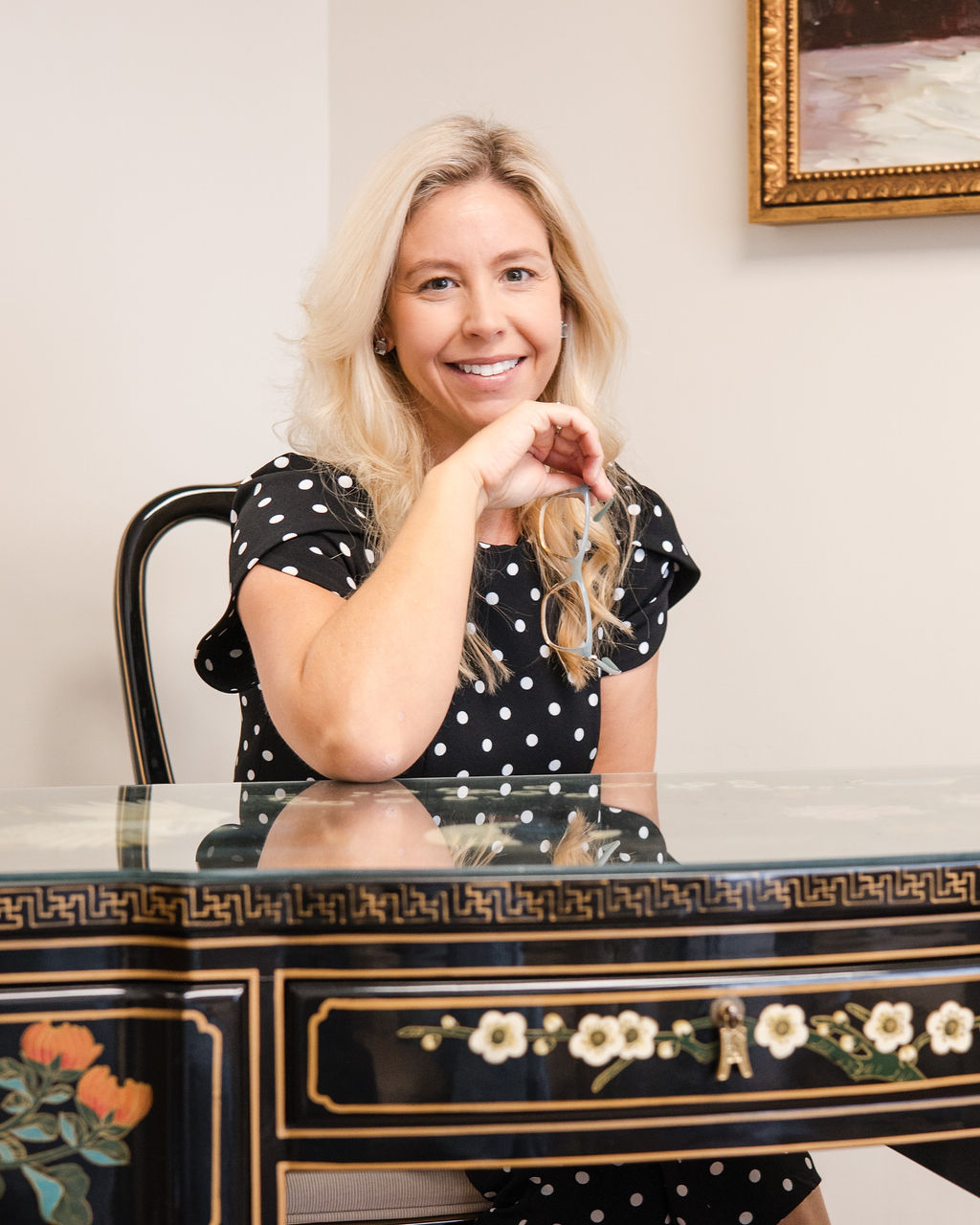 Lindsey Capelli, Living Well Counseling Center owner, in a black polka dot dress sitting at a decorative glass-top table holding eyeglasses.