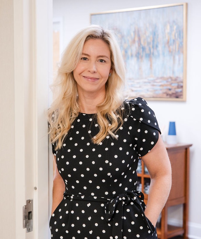Lindsey Capelli, owner of Living Well Counseling Center, and Licensed Therapist in a black and white polka dot dress sitting at a decorative black table with floral designs holding eyeglasses.