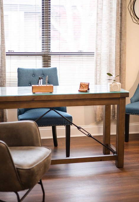 A cozy corner in the therapy practice with a blue armchair, textured pillow, wooden electric fireplace, and a floor lamp near window with sheer curtains.