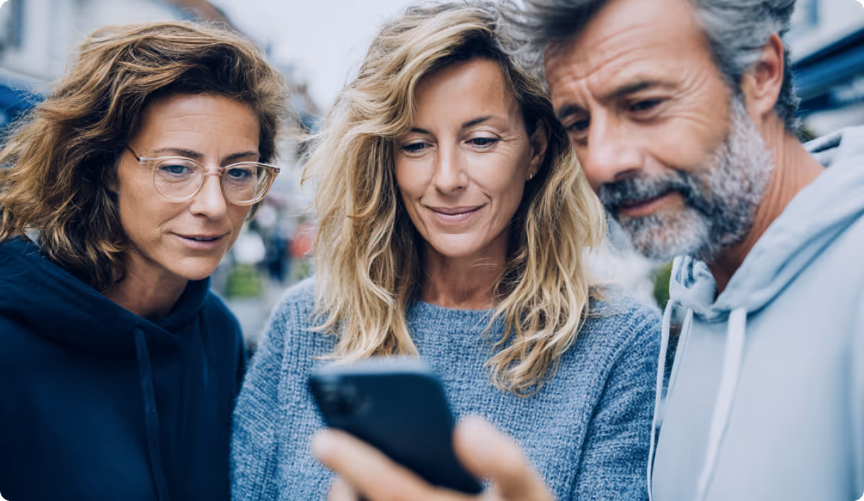 Group of people looking at a smartphone - Three people—two women and one man—gathered closely together, looking down at a smartphone screen with engaged expressions.
