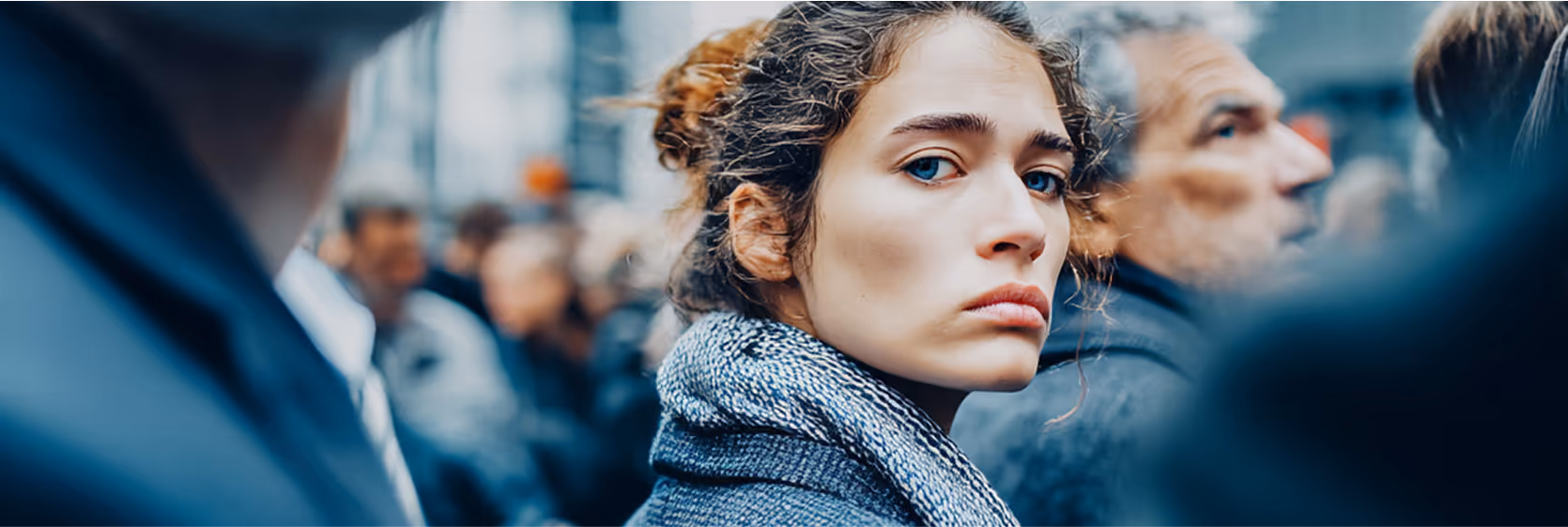 Woman looking thoughtfully in a crowd - A close-up of a woman with curly hair looking to the side with a contemplative expression, with blurred people in the background.