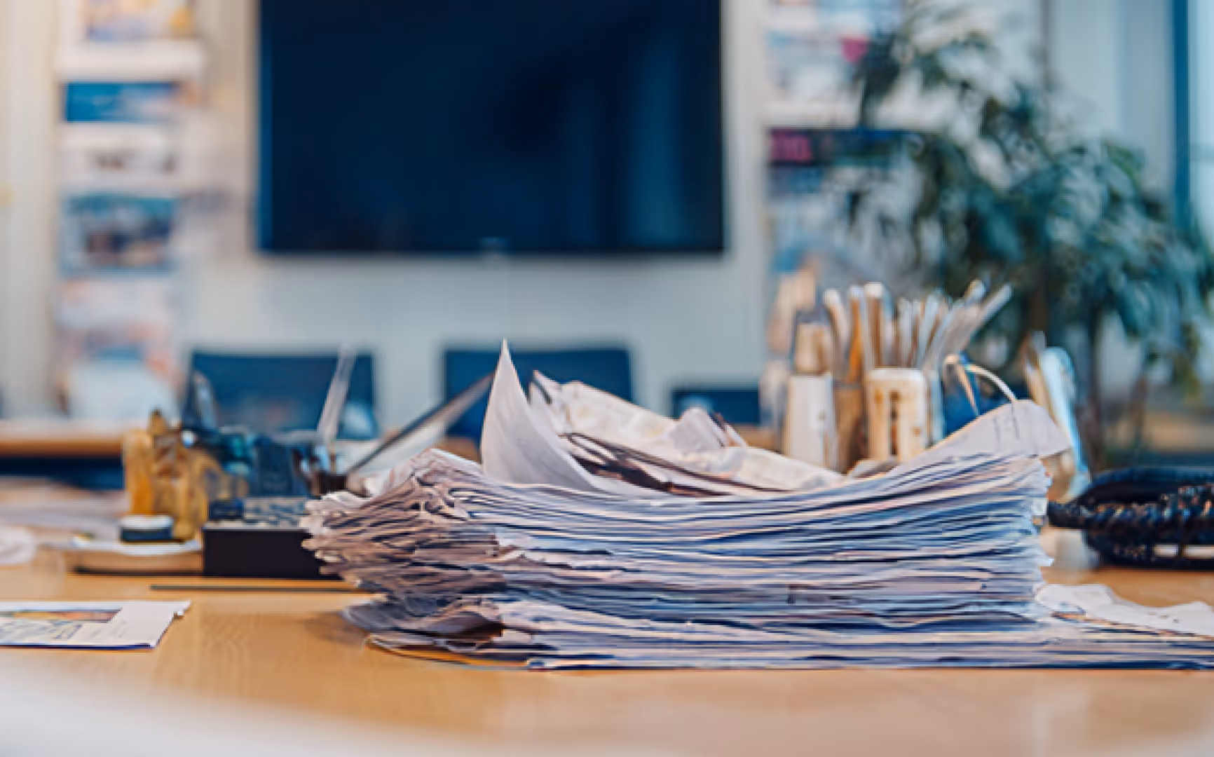 Stack of papers on a desk - A large pile of documents or papers sits on a desk, with a blurred office background featuring a computer monitor and a potted plant.
