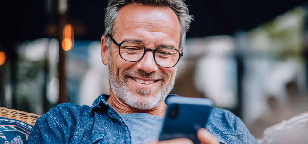 Smiling older man with glasses wearing a blue denim shirt, photographed in casual setting with soft background lighting