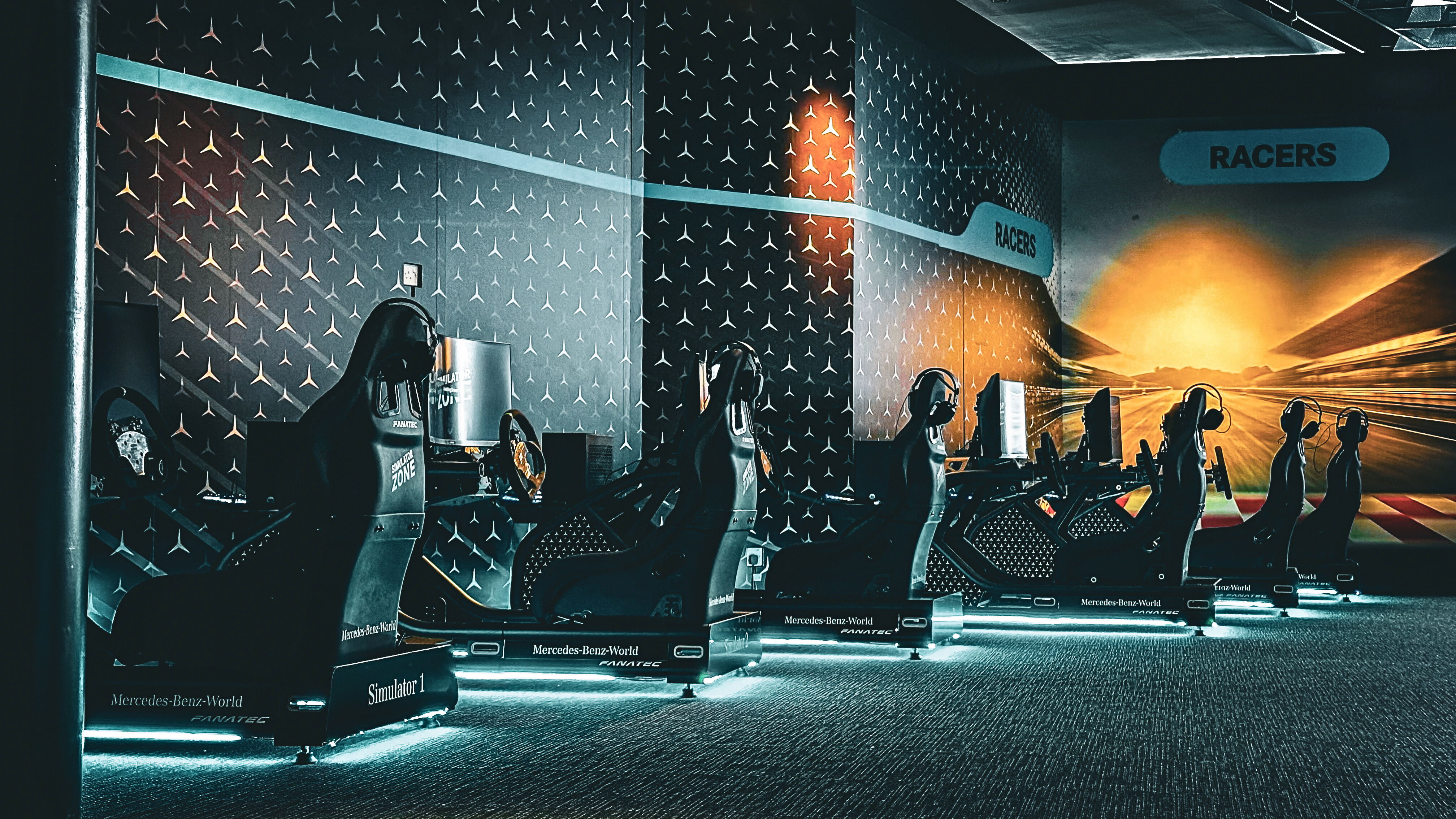 Row of black racing simulators with steering wheels and monitors in a dark gaming room.