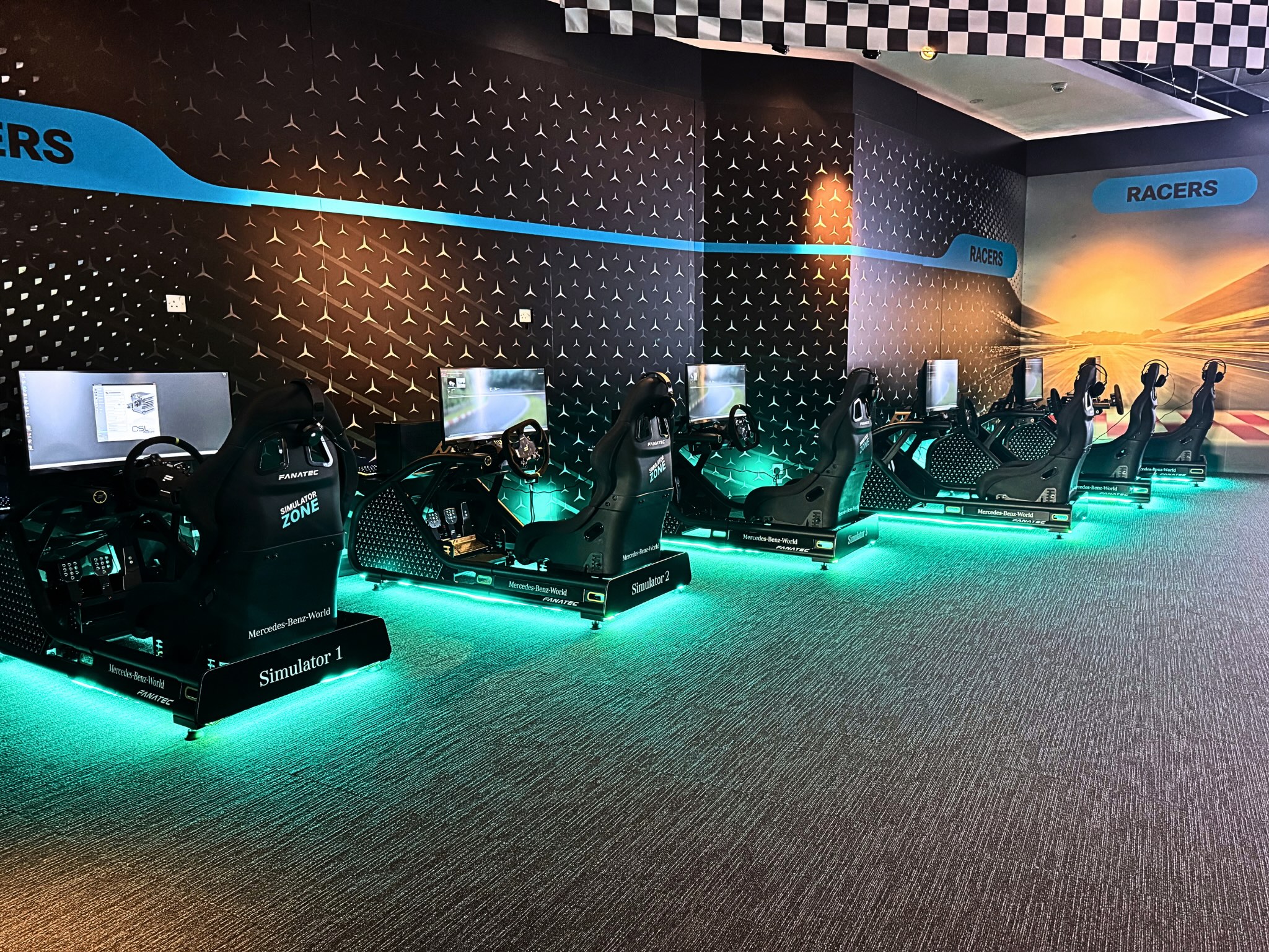 Row of seven racing simulators with black seats, steering wheels, and screens in a themed room.
