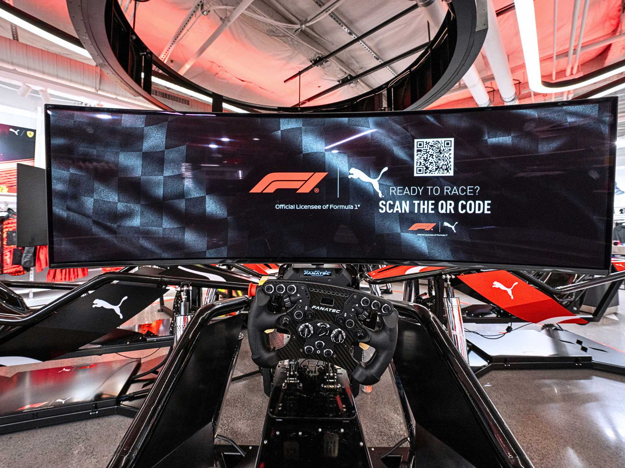 Sim racing setup with a Fanatec steering wheel and a curved screen showing a Puma and F1 QR code promo.