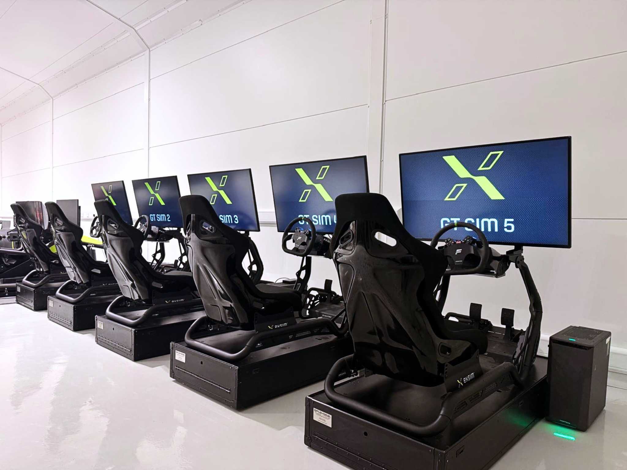 Row of five black racing simulator seats with steering wheels and monitors displaying GT Sim 1 to 5.