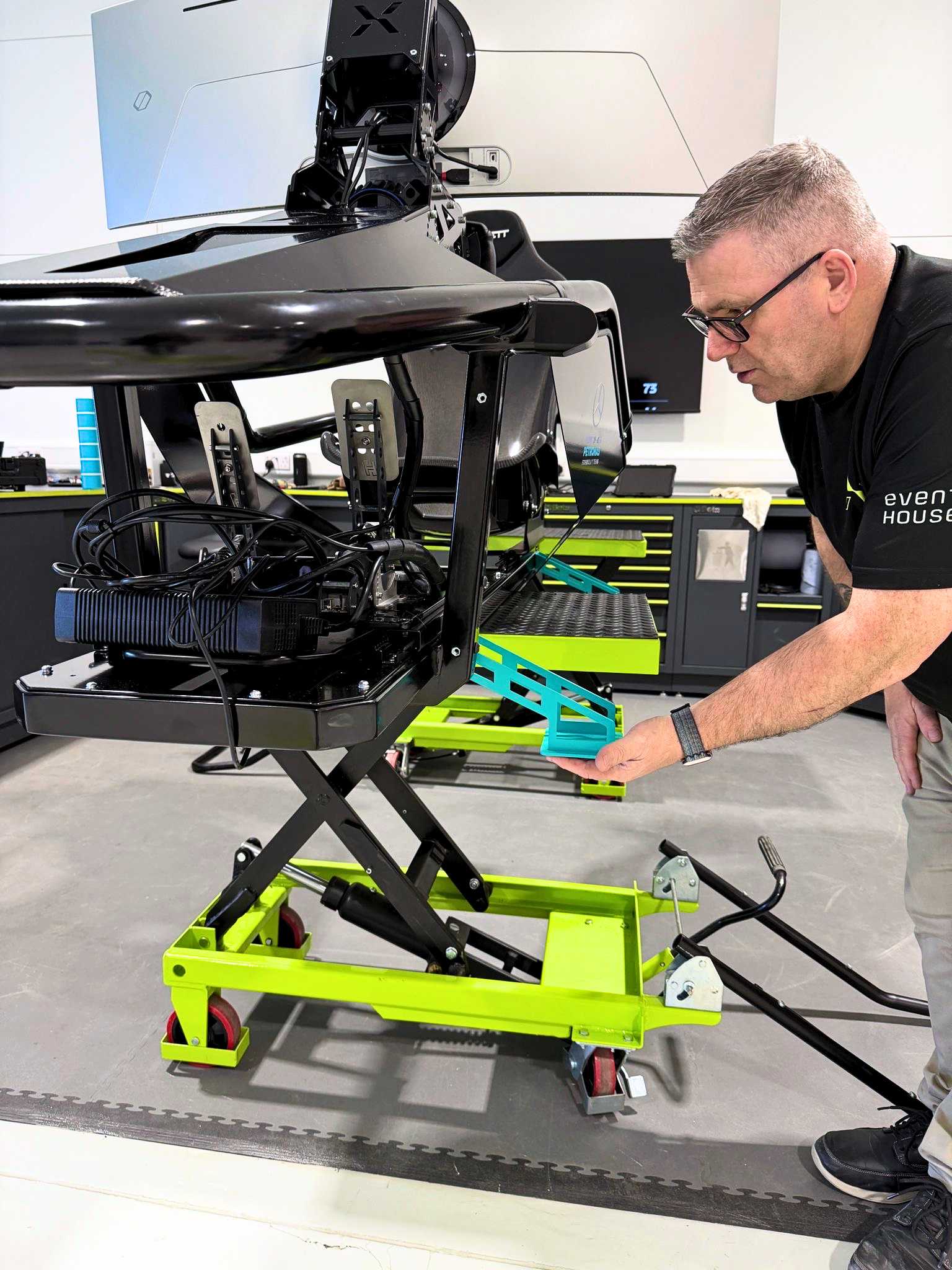 Man adjusting a simulator platform with pedals, lifted on a green hydraulic stand in a workshop.