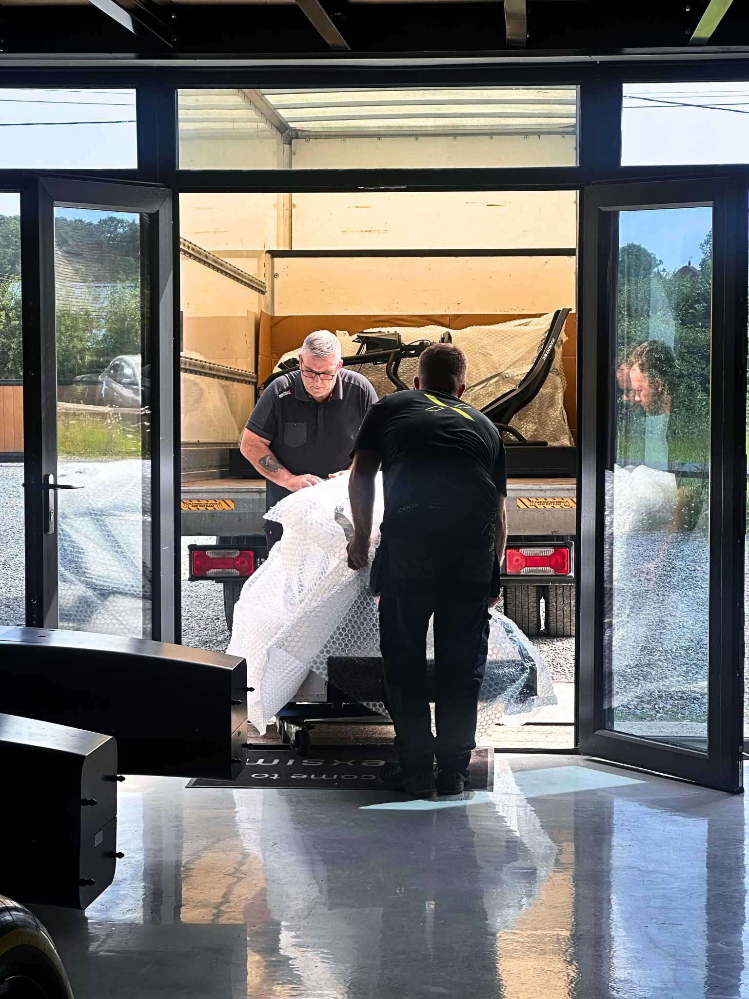 Two men loading a bubble-wrapped item from a truck into a building through open glass doors.