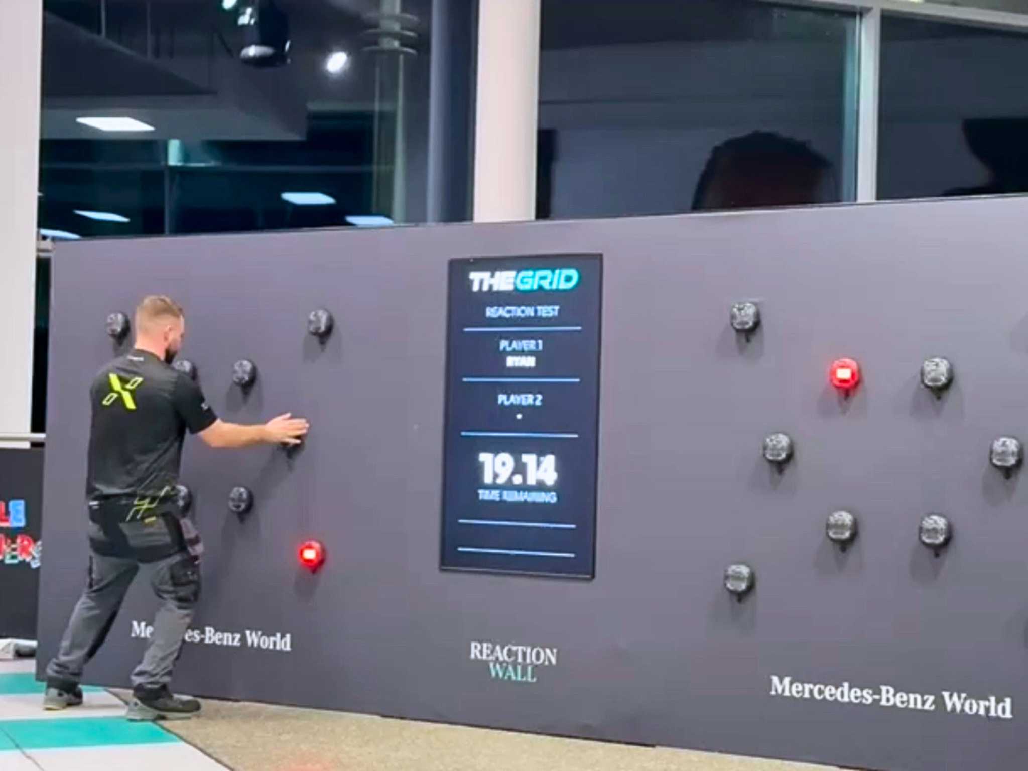 Man in black reacts to lights on a gray reaction test wall with timer display in a Mercedes-Benz World facility.