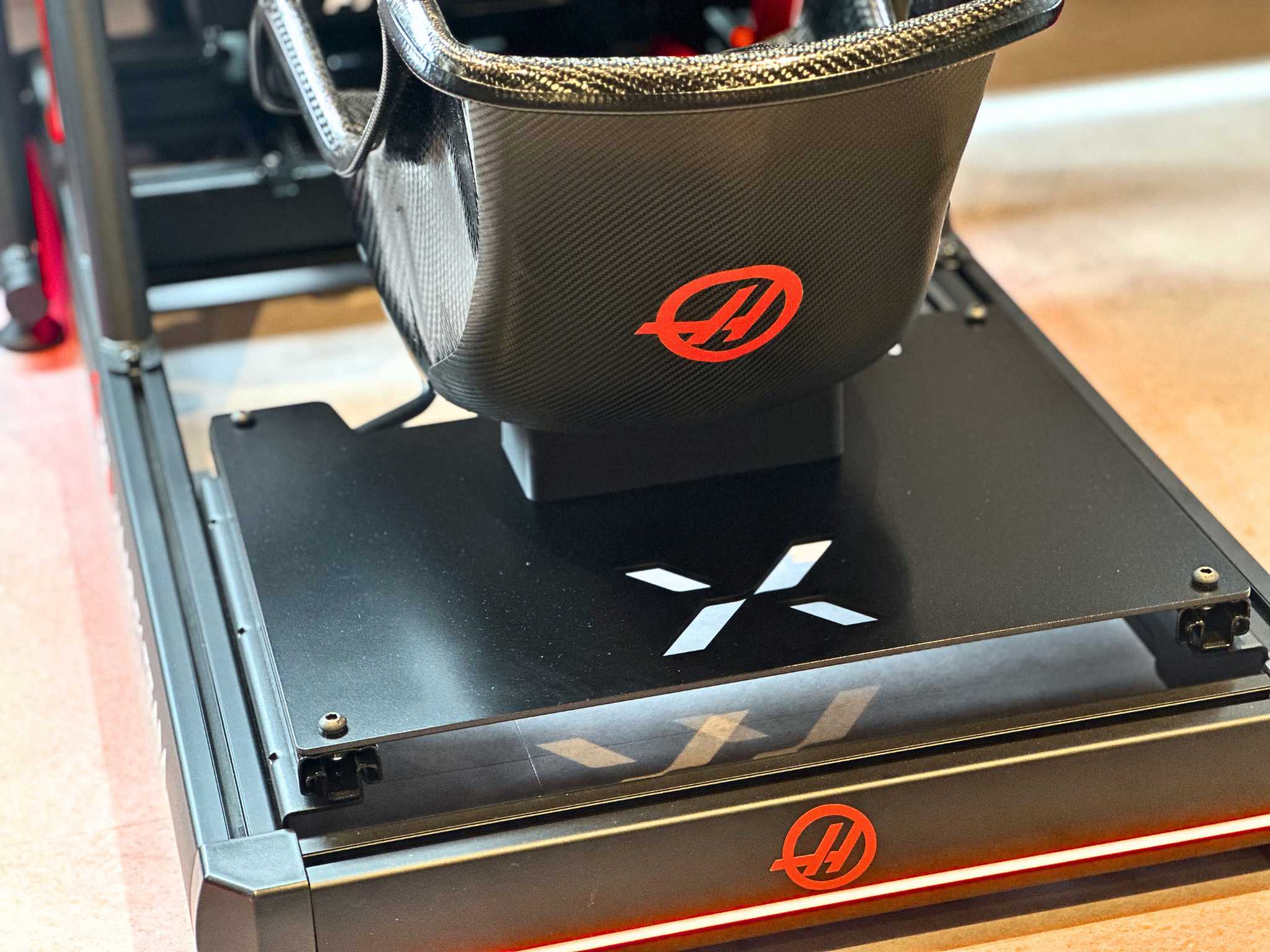 Close-up of a black carbon fiber racing simulator seat with red logo on black base and platform.