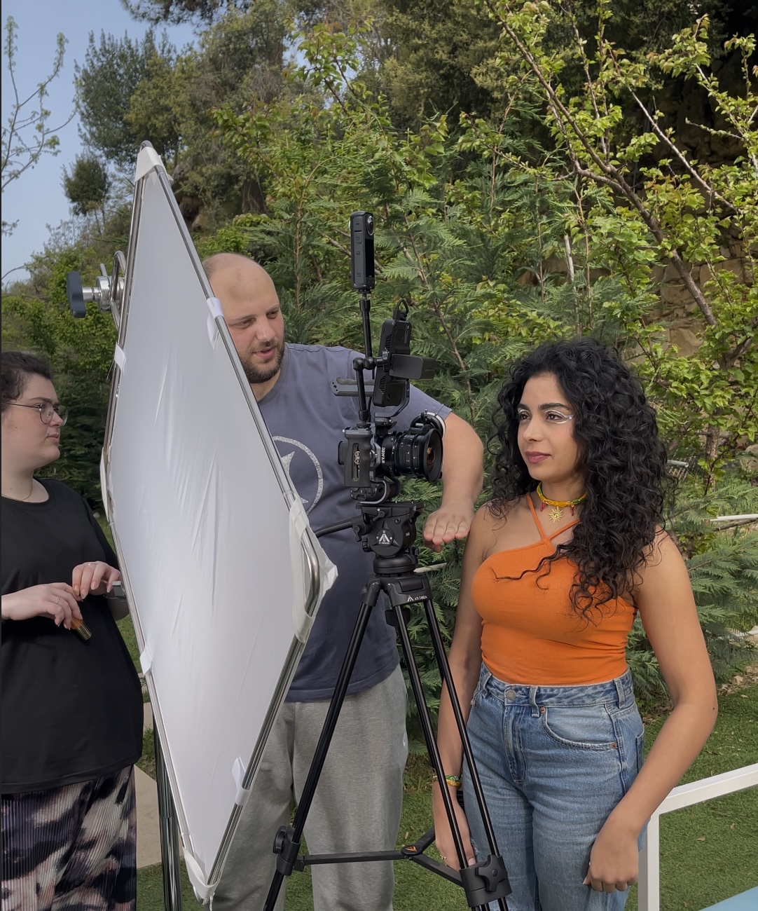 Three people outdoors with camera equipment and a reflector screen set up in a green area with trees.| MMNT PRODUCTIONS