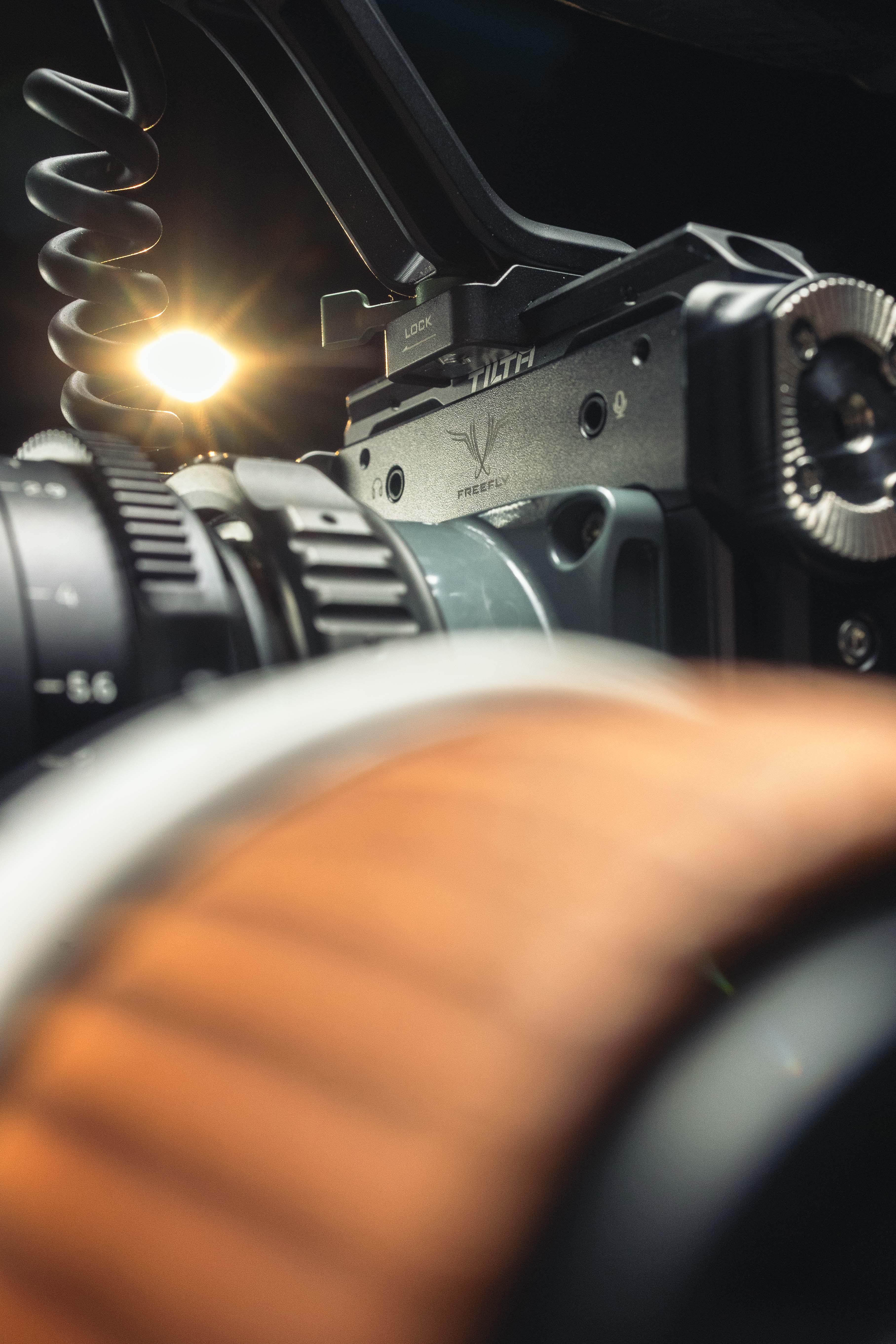 Close-up of a professional camera rig with lens gears, wooden handle, and a coiled cable under bright lighting.| MMNT PRODUCTIONS