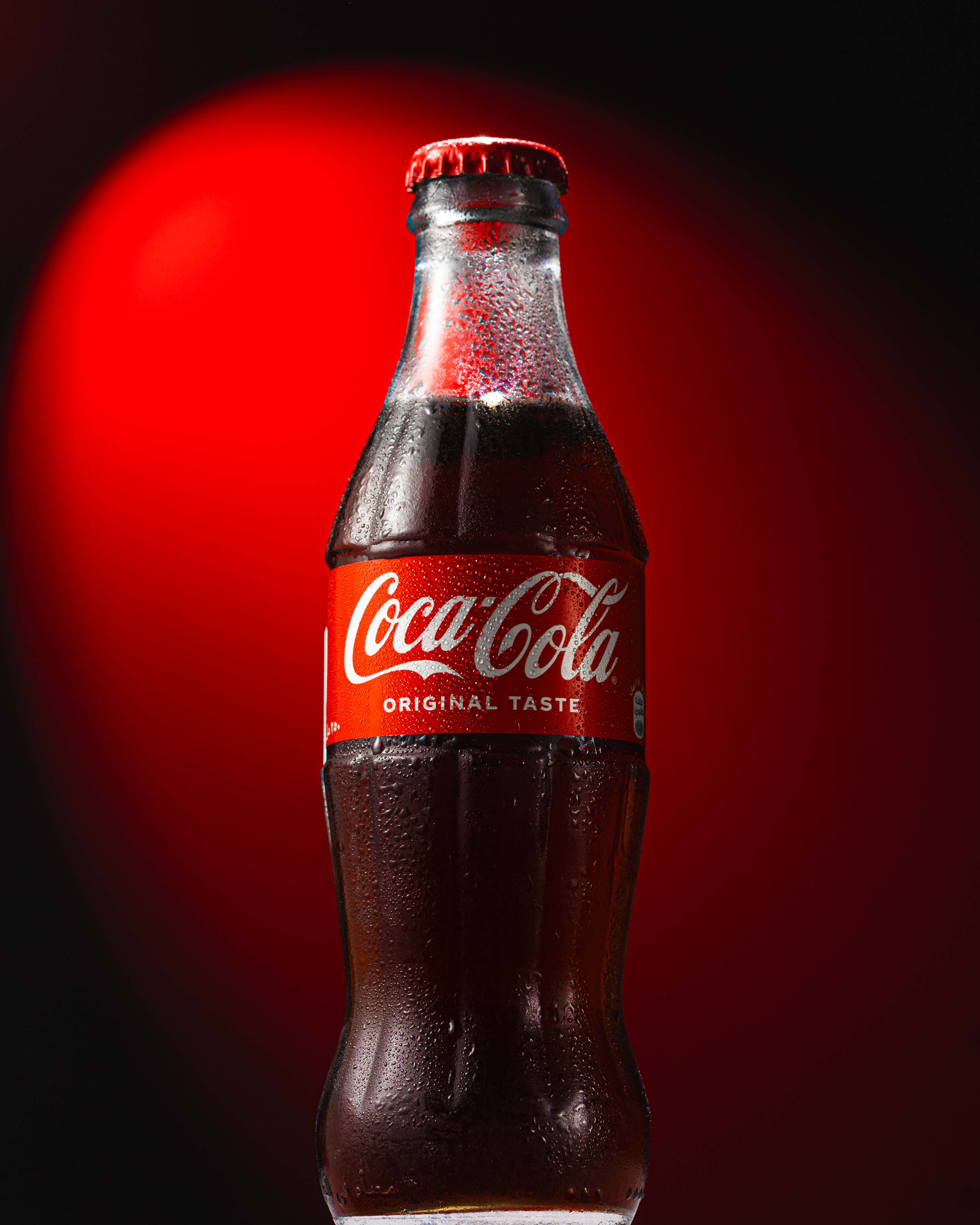Cold glass bottle of Coca-Cola with condensation against a red and black background.| MMNT Productions
