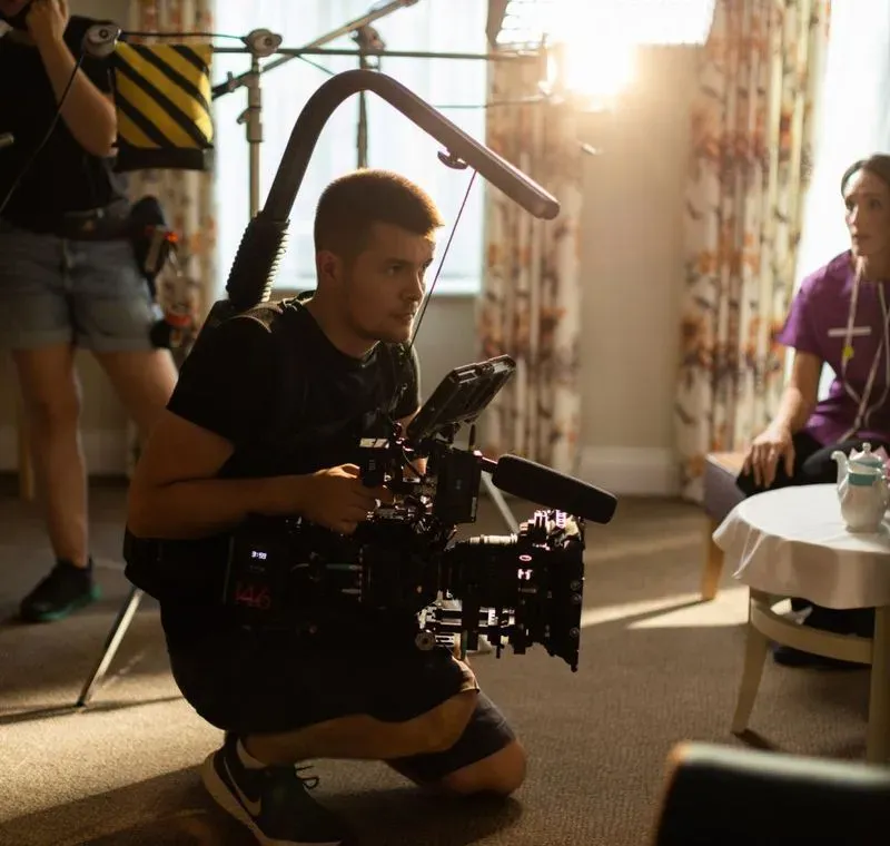 Cameraman films a nurse for a scene, with natural light and set props in view.