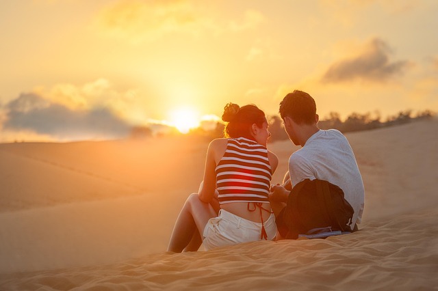 Couple assis sur une dune de sable, observant un magnifique coucher de soleil sur un paysage désertique.