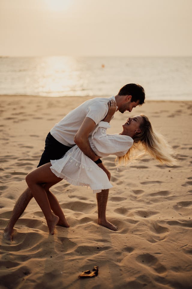 Jeune couple riant et dansant sur une plage de sable au coucher du soleil, profitant de leurs vacances