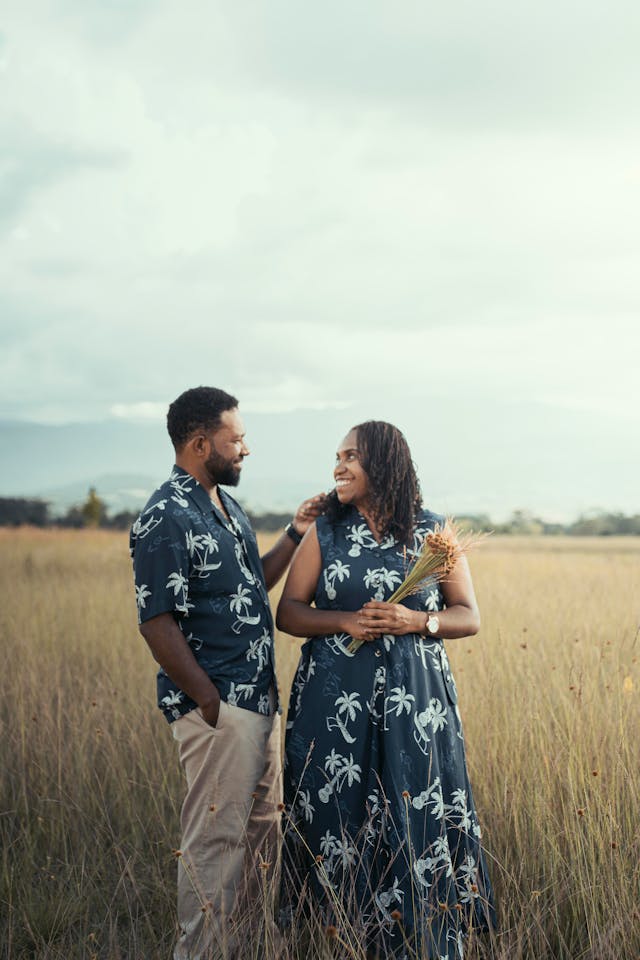 Couple souriant et heureux posant dans un champ d'herbes hautes, l'homme en chemise à motifs et la femme tenant des fleurs sèches.