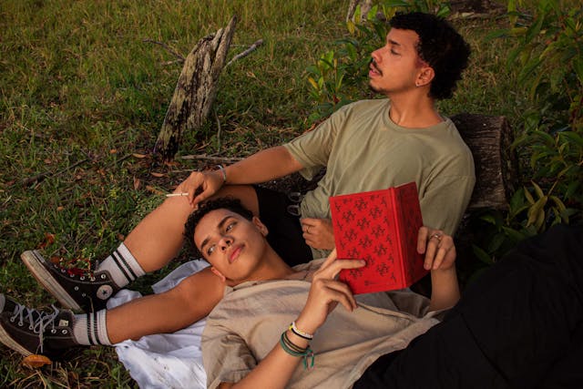 Deux hommes se relaxant dans l'herbe, l'un lisant un livre rouge, dans un cadre naturel paisible.