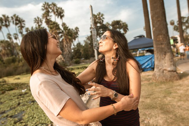 Deux femmes souriantes se tenant et riant sous le soleil, avec des palmiers en arrière-plan, en plein air.