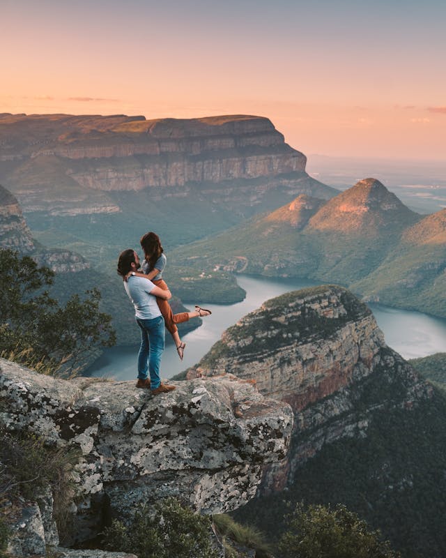 Couple s'embrassant sur un rocher avec un paysage de montagnes et de rivière spectaculaire en arrière-plan.